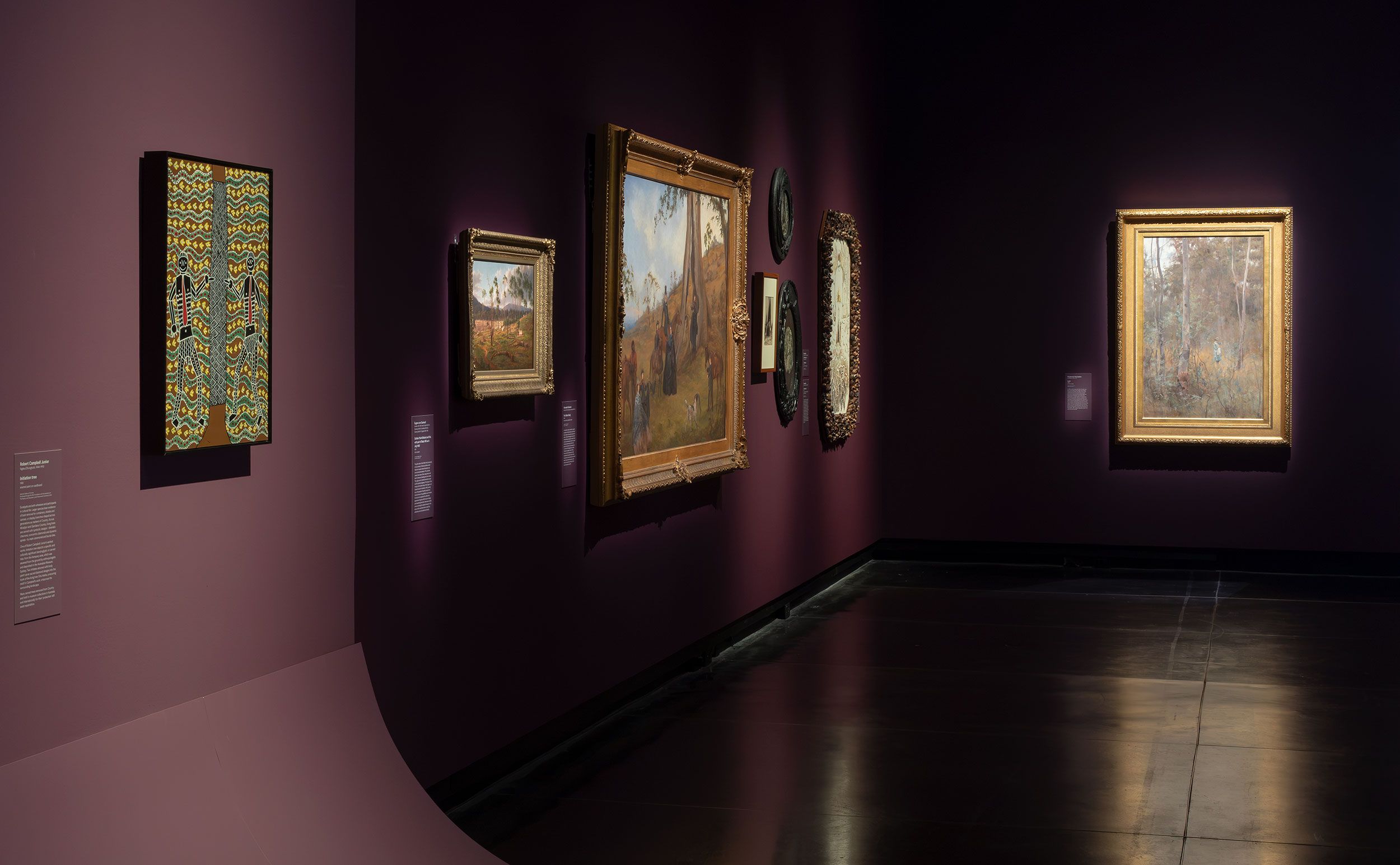 Art gallery room with dark purple walls displaying various framed paintings and artworks, illuminated by spotlights. The artworks vary in size and style, and museum labels are visible next to each piece.