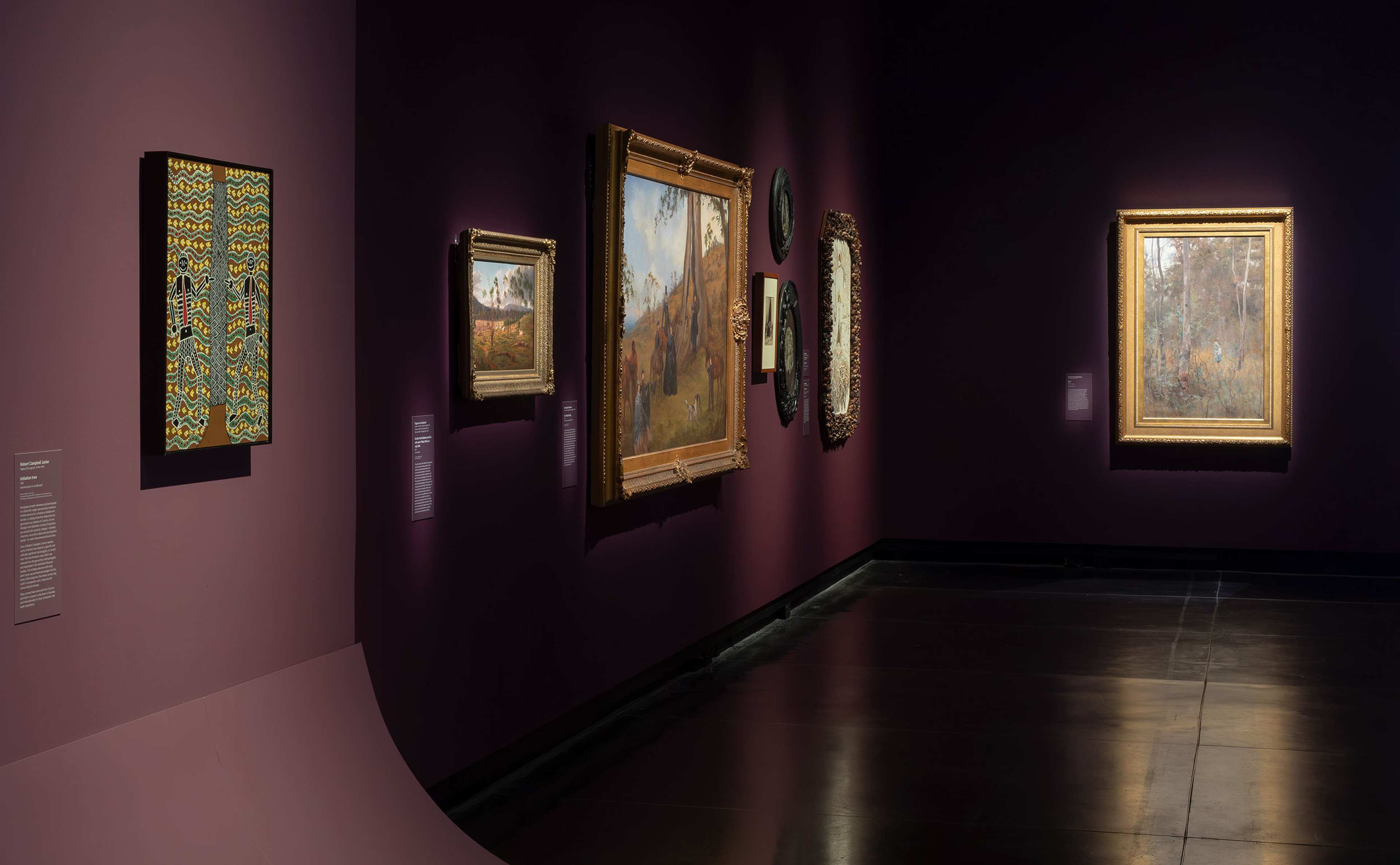 Art gallery room with dark purple walls displaying various framed paintings and artworks, illuminated by spotlights. The artworks vary in size and style, and museum labels are visible next to each piece.
