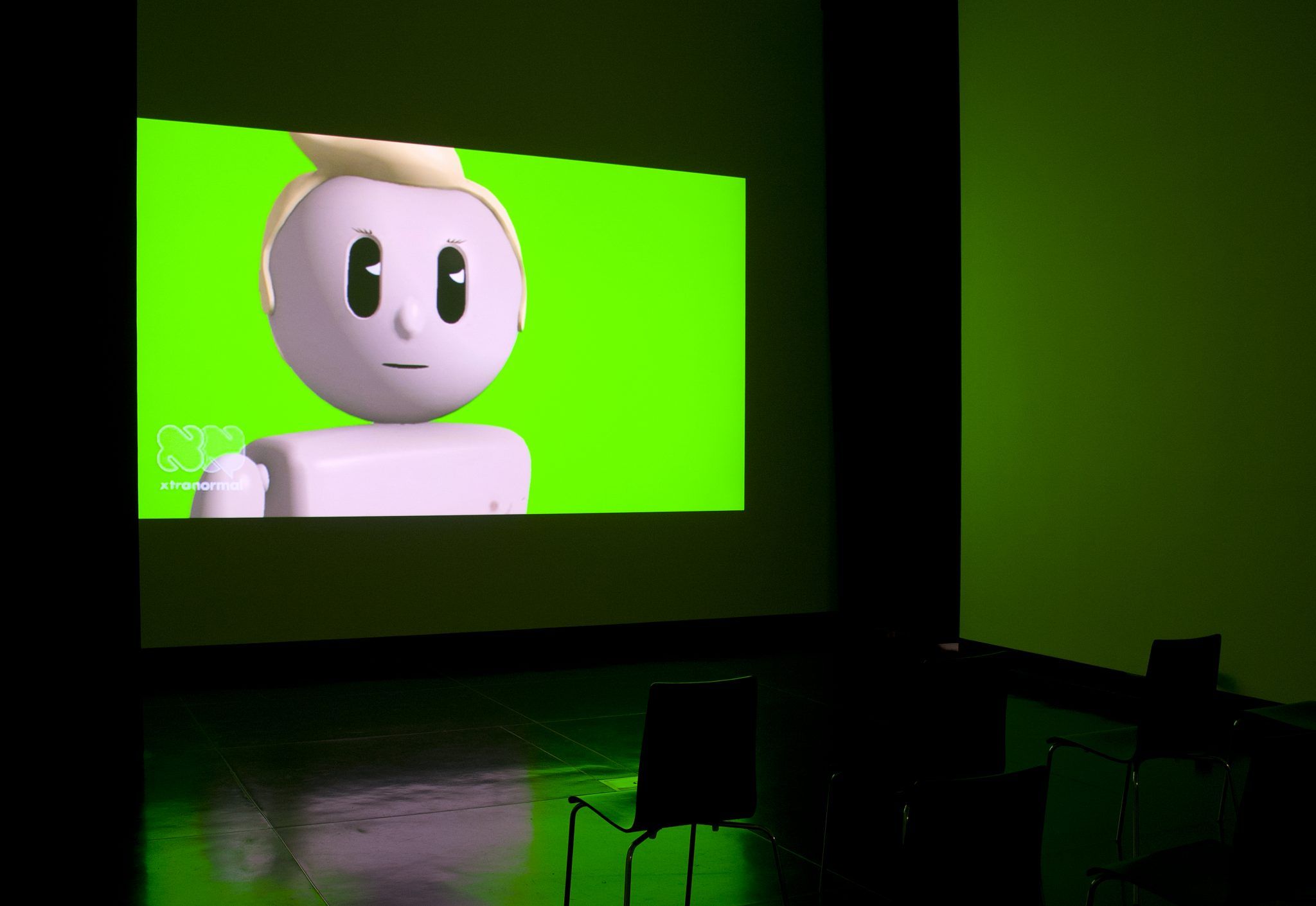 A photograph of a darkened space with a chair in front of a projection of an animated video with a lego-like figure against the flour green background
