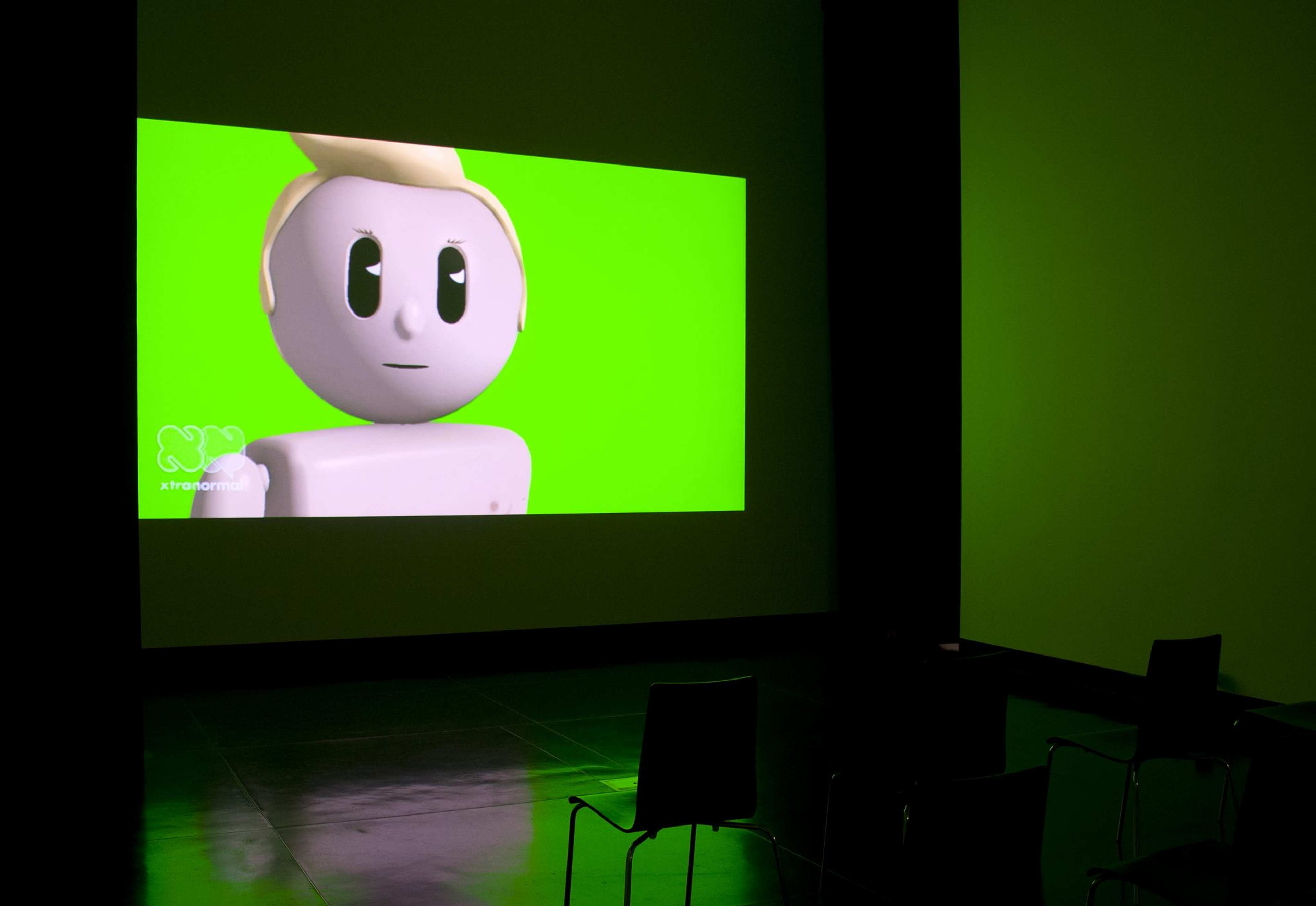 A photograph of a darkened space with a chair in front of a projection of an animated video with a lego-like figure against the flour green background