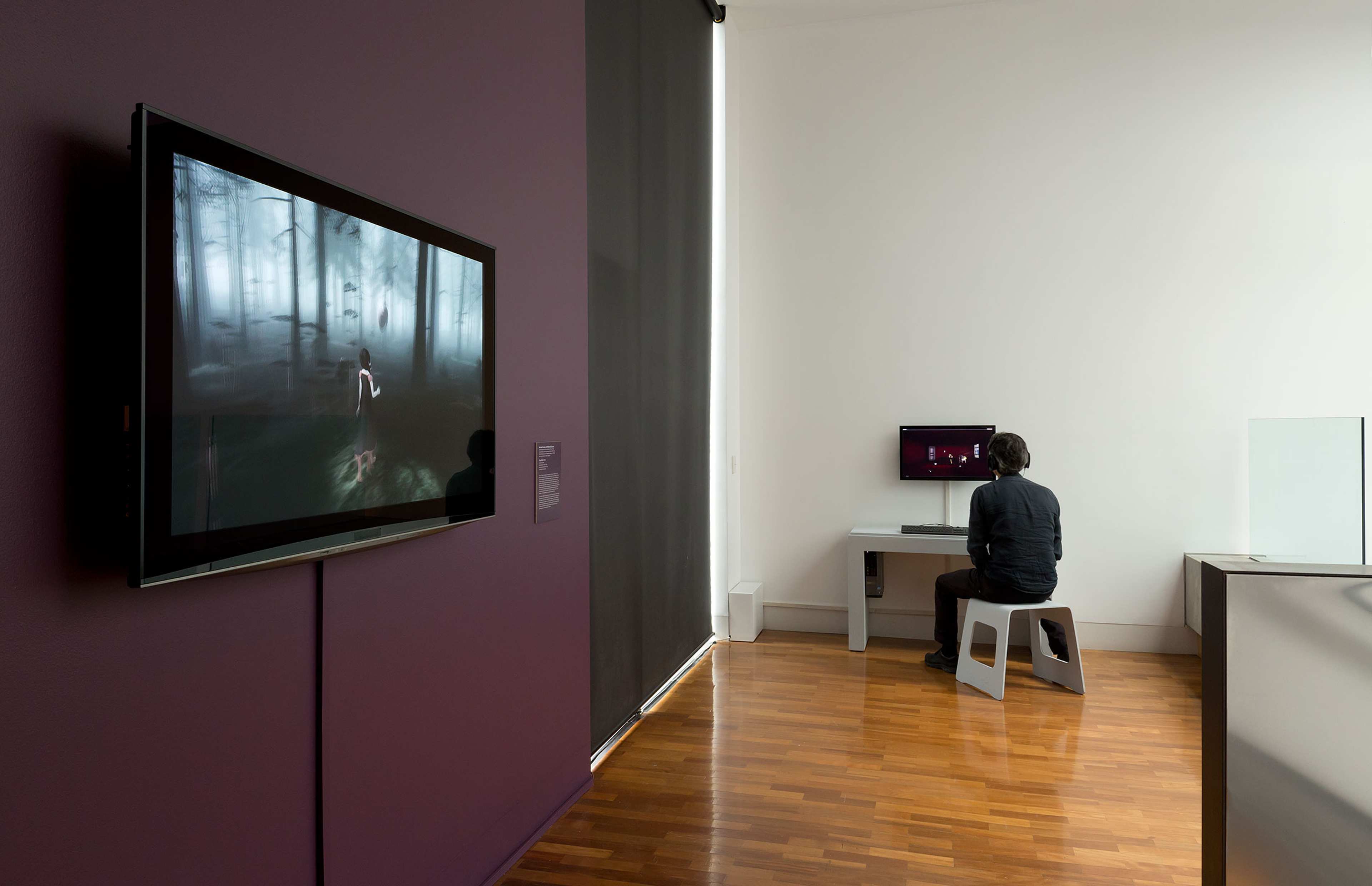 A person sits on a stool using a computer in a modern, minimal room with wooden floors. On the left wall, a screen displays a dark, misty forest scene from a video game.