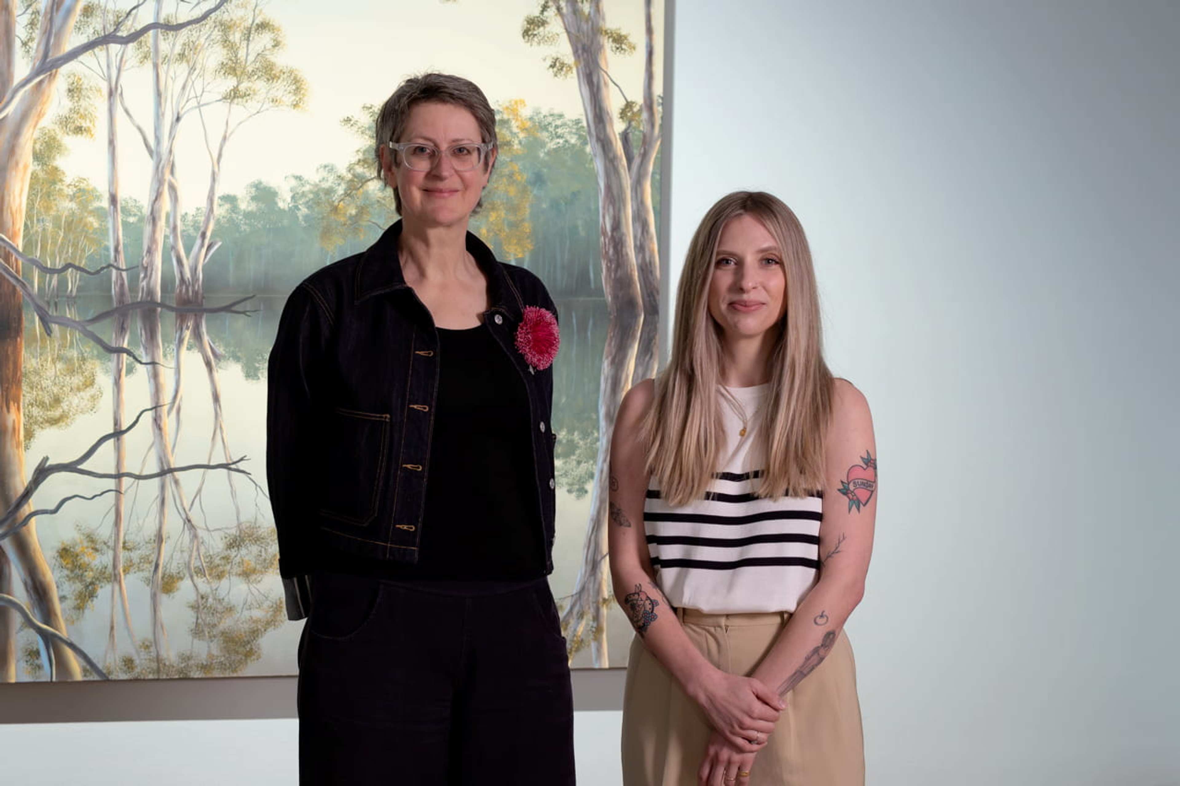 Two women are shown side by side against a painting of a gum tree in water. The woman on the left has short hair, glasses, and wears a black suit. The woman on the right has long blonde hair and is standing with her hands clasped.