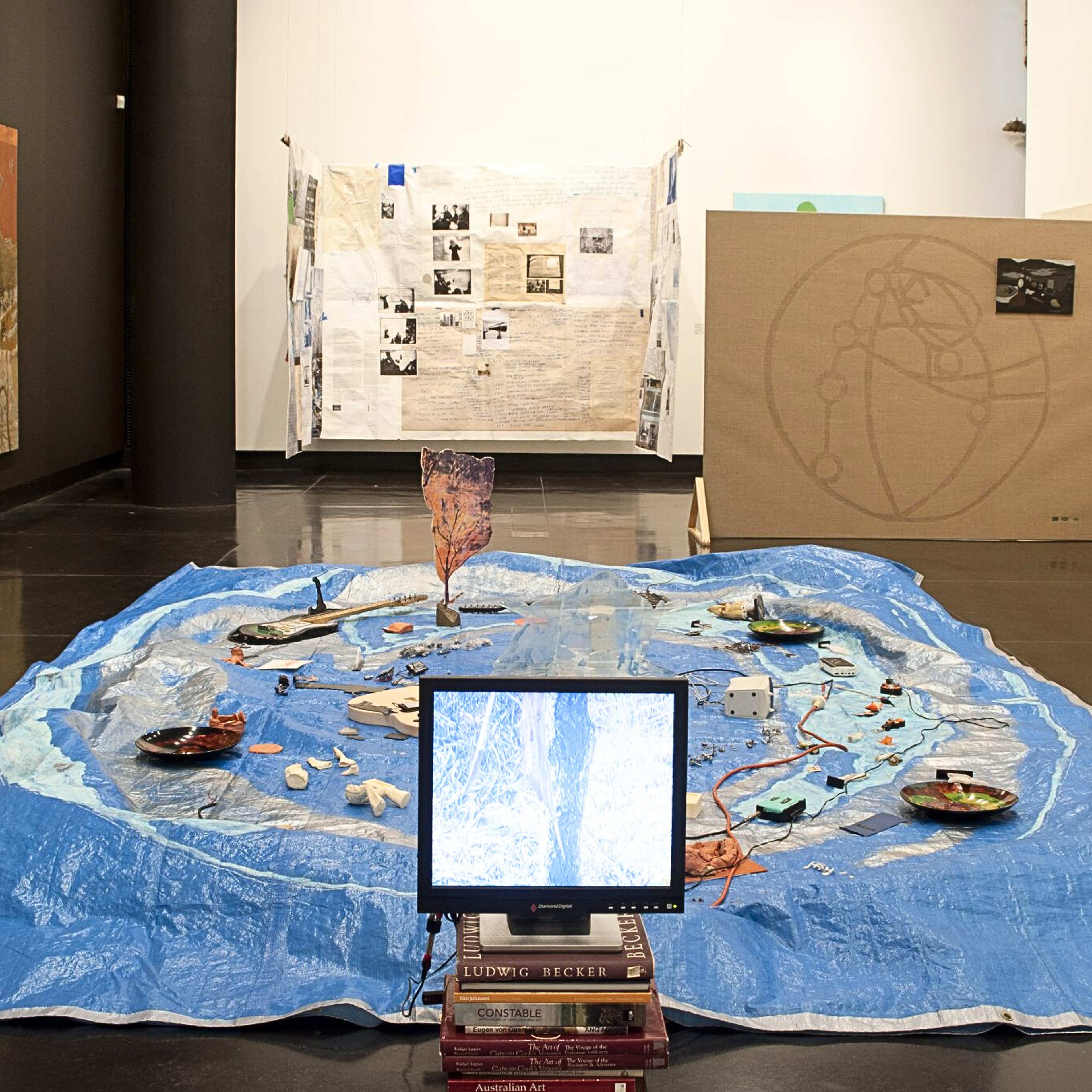 A photograph of an exhibition space with a tarp with a collection of objects occupying most of the floor, a computer display in front of it and large format drawings and assemblages on the walls around