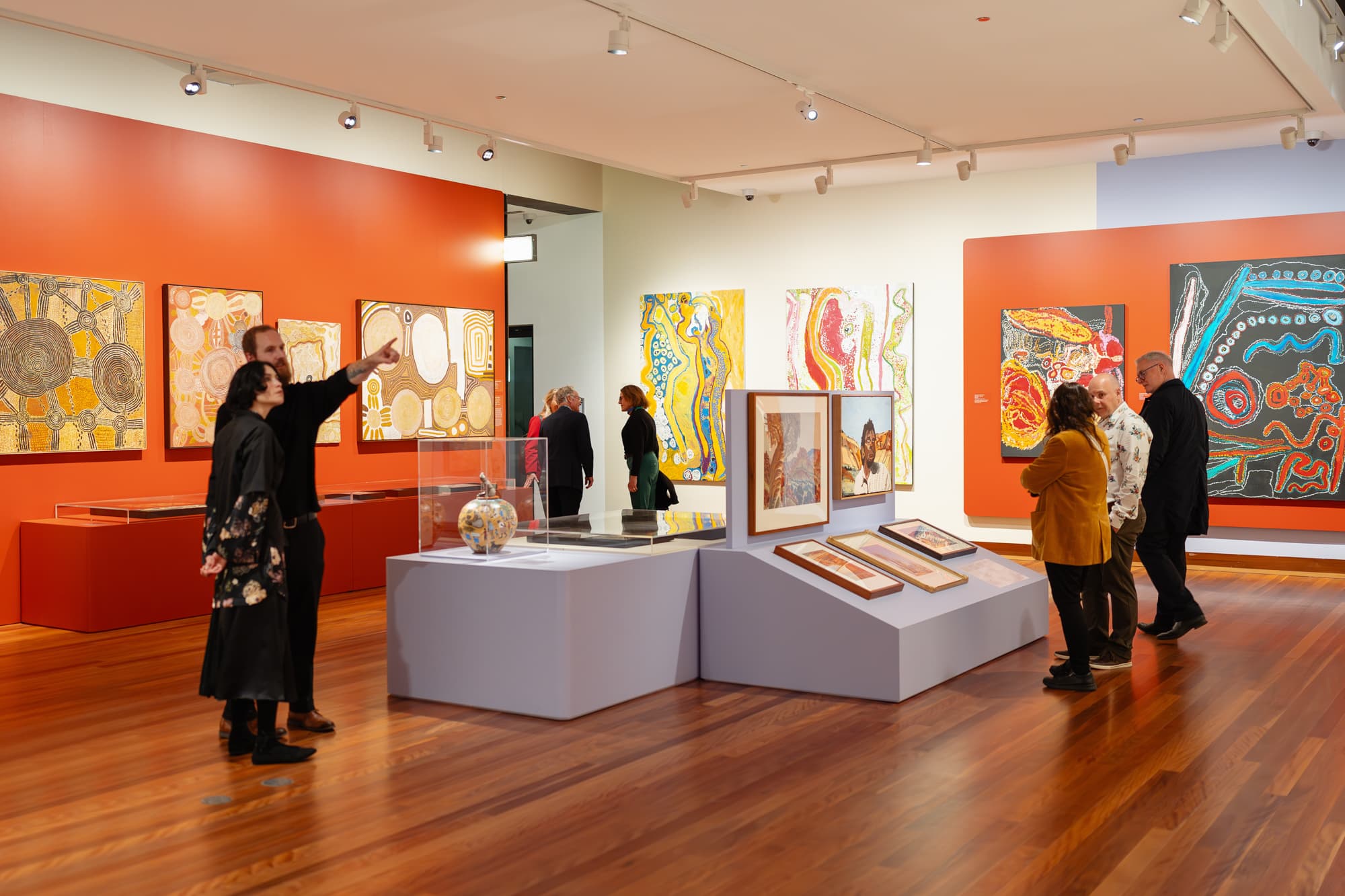 Visitors view and discuss colourful abstract artworks and artefacts displayed in a modern art gallery with red accent walls and wood floors. Some people read information panels, while others observe the exhibits together.