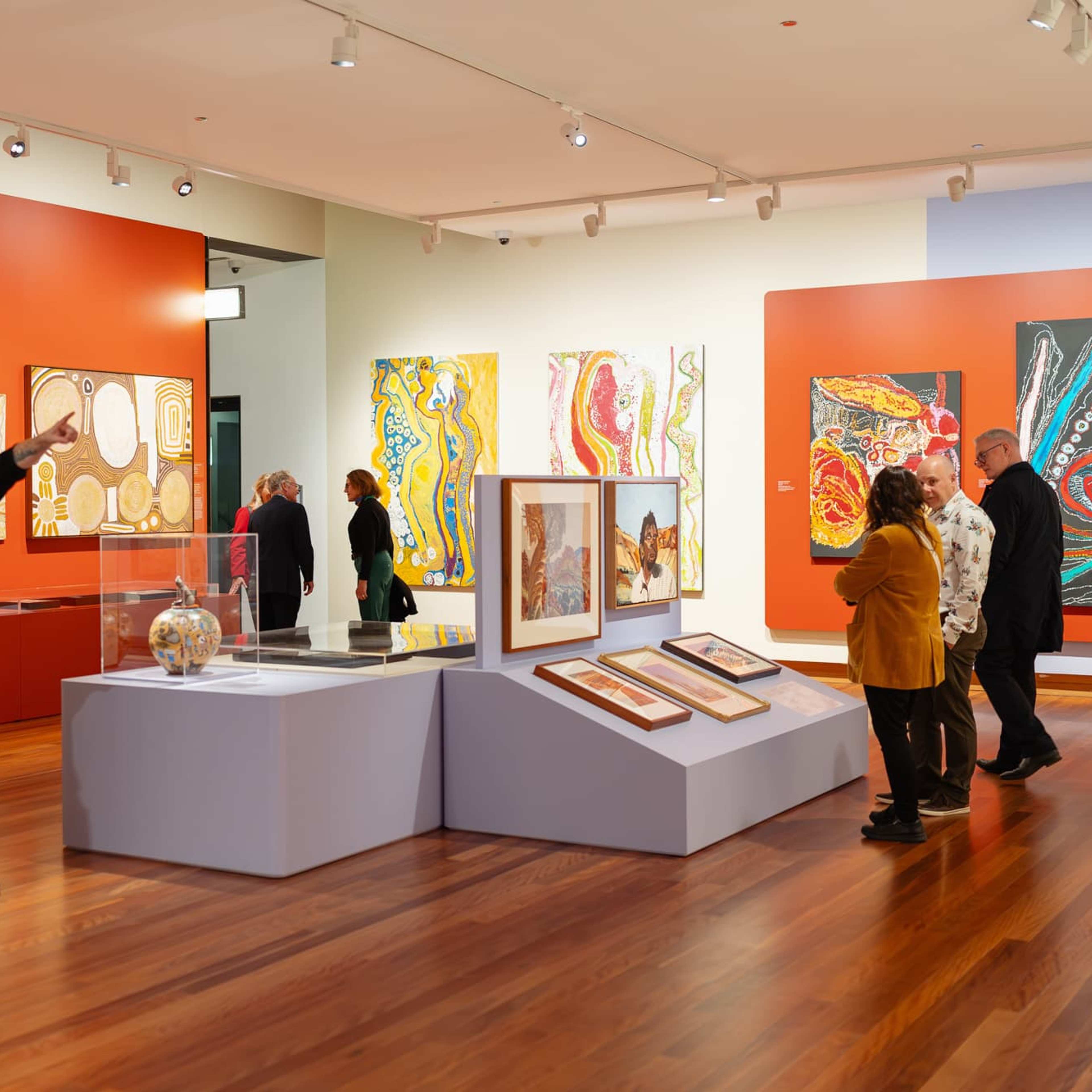 Visitors view and discuss colourful abstract artworks and artefacts displayed in a modern art gallery with red accent walls and wood floors. Some people read information panels, while others observe the exhibits together.