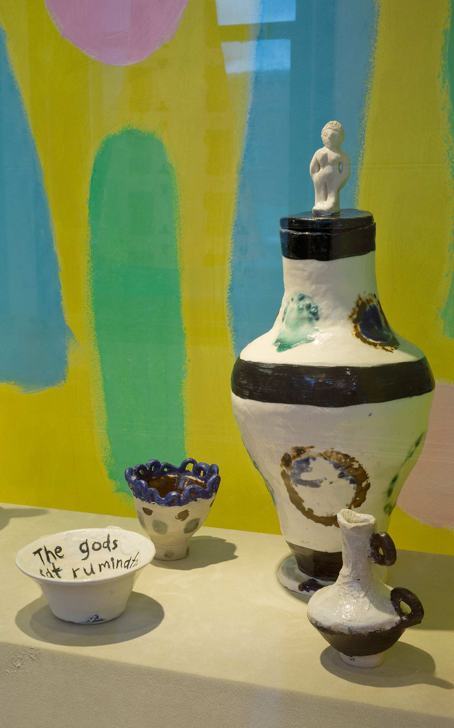 A photograph of contemporary black and white ceramic bowls and vases arranged on a plinths against a colourful drawn background featuring yellow and light blue and green
