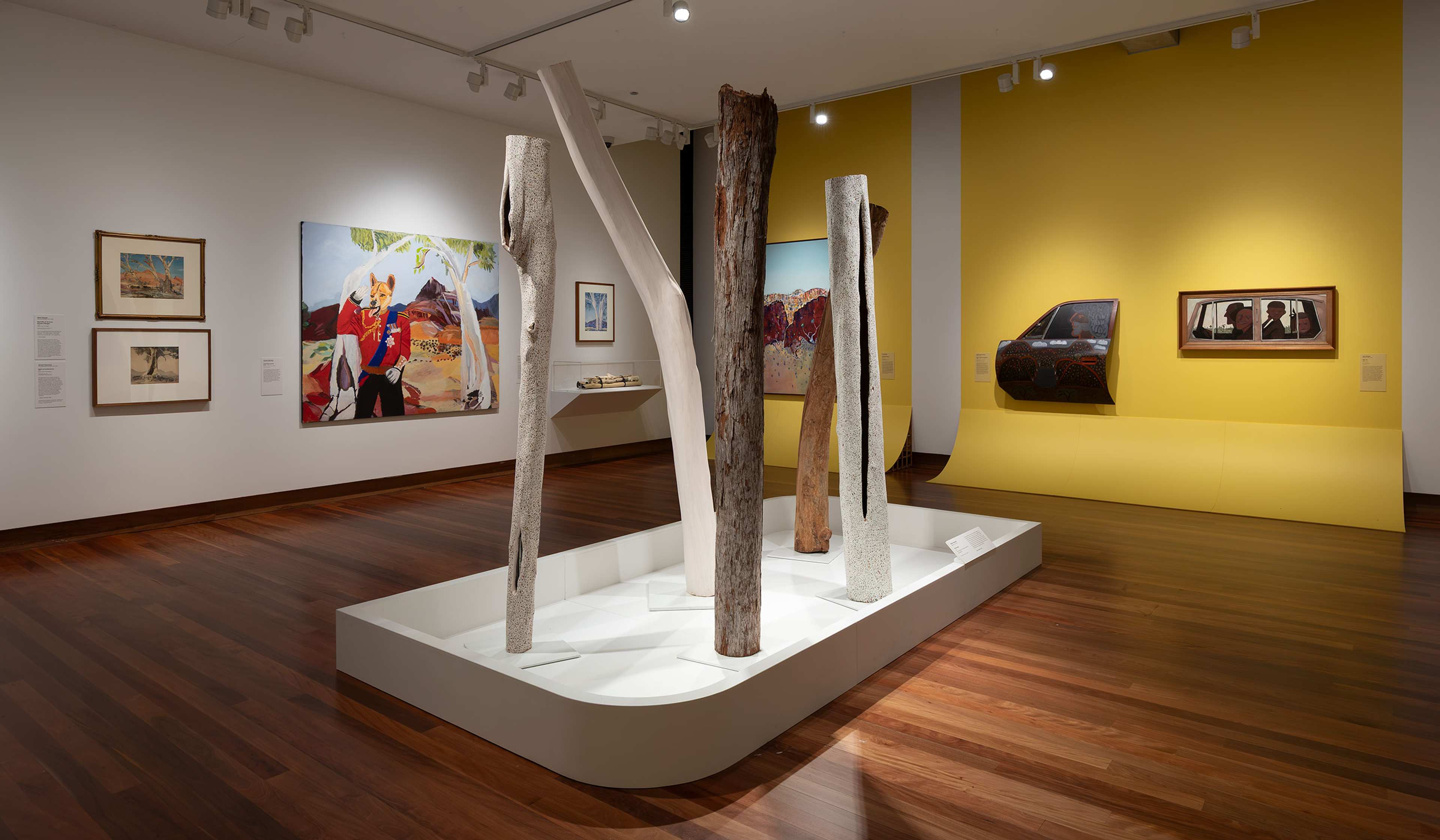 A modern art gallery with polished wooden floors, featuring tall tree trunk sculptures in the center, colorful paintings on the walls, and a bright yellow accent wall in the background.