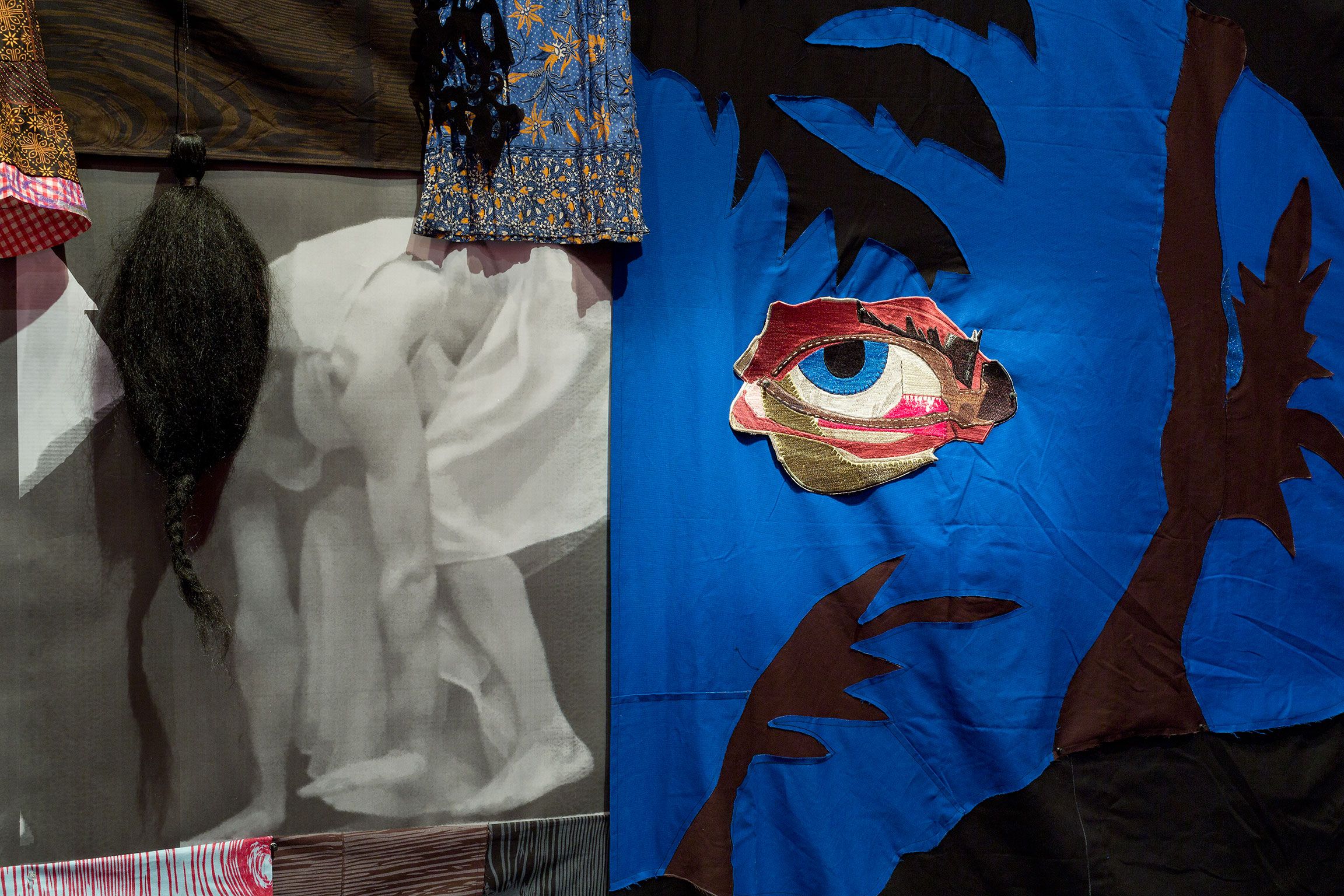 A close-up photograph of a fabric collage with a figure bent over and wig attached on the left side and a cutout of an eye on blue background in the right half