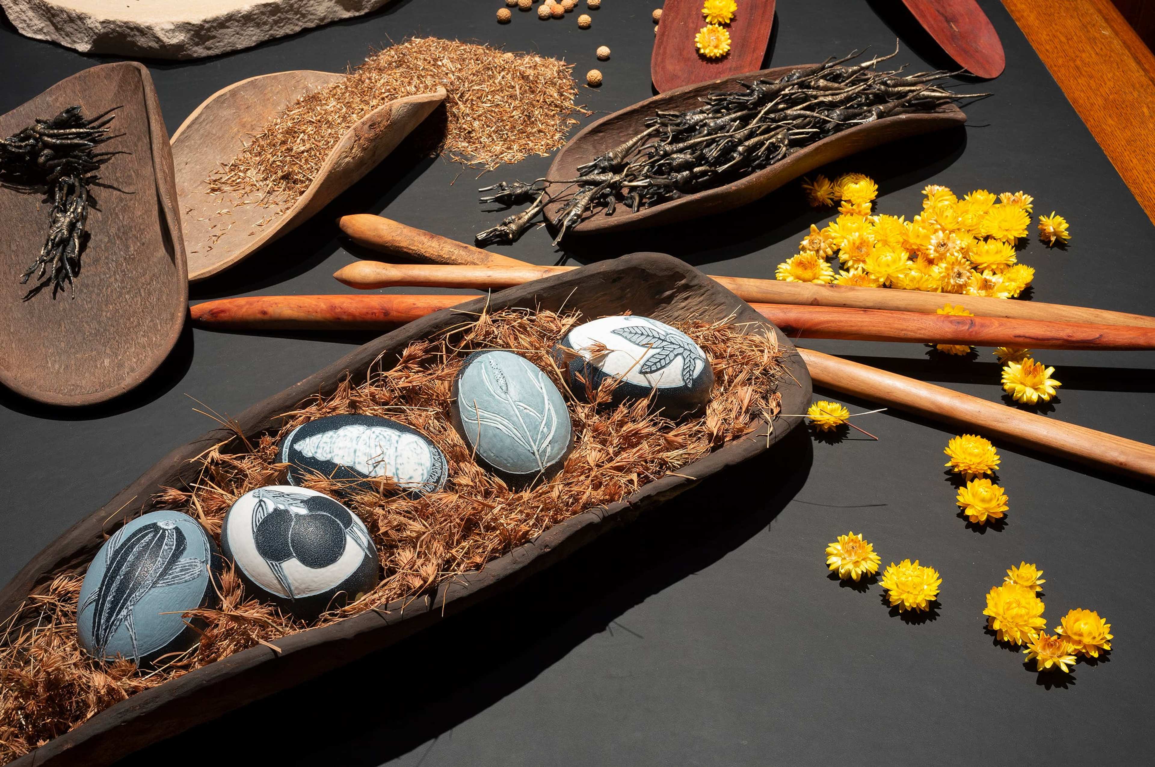 Decorated eggs with intricate designs rest on dried grass in a wooden tray, surrounded by yellow flowers, twigs, seeds, and wooden utensils on a dark surface.