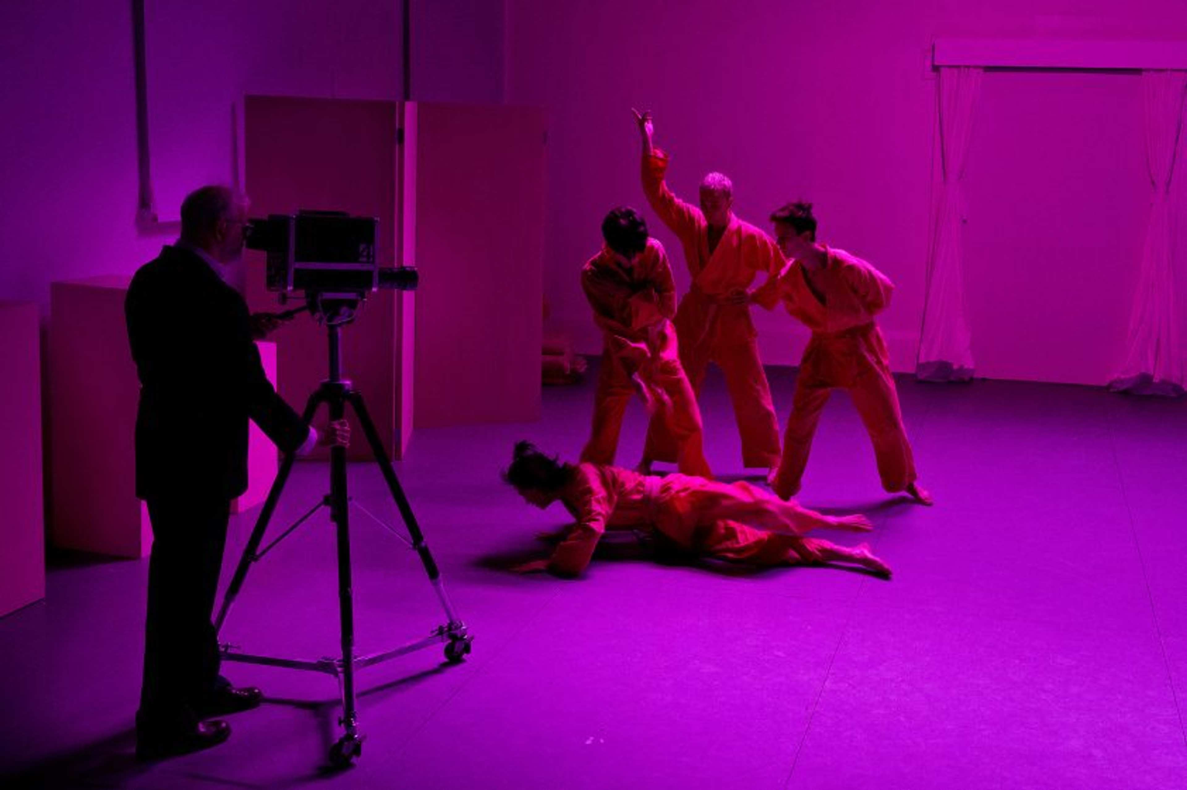 A photographer with a camera on a tripod takes pictures of four people in red outfits performing expressive poses under purple lighting on a stage. One person is on the floor, while three others stand, gesturing dramatically.
