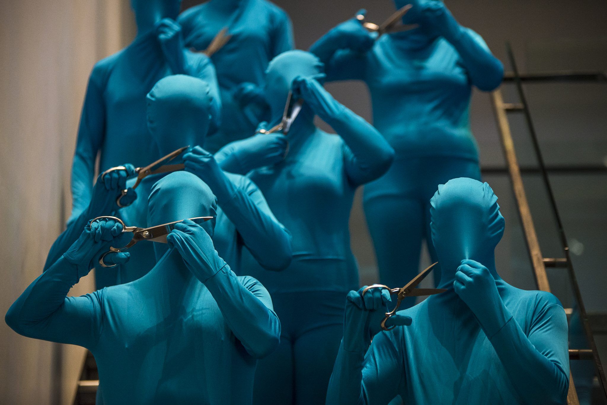 Eight performers in blue body suits use scissors to cut a hole in the fabric