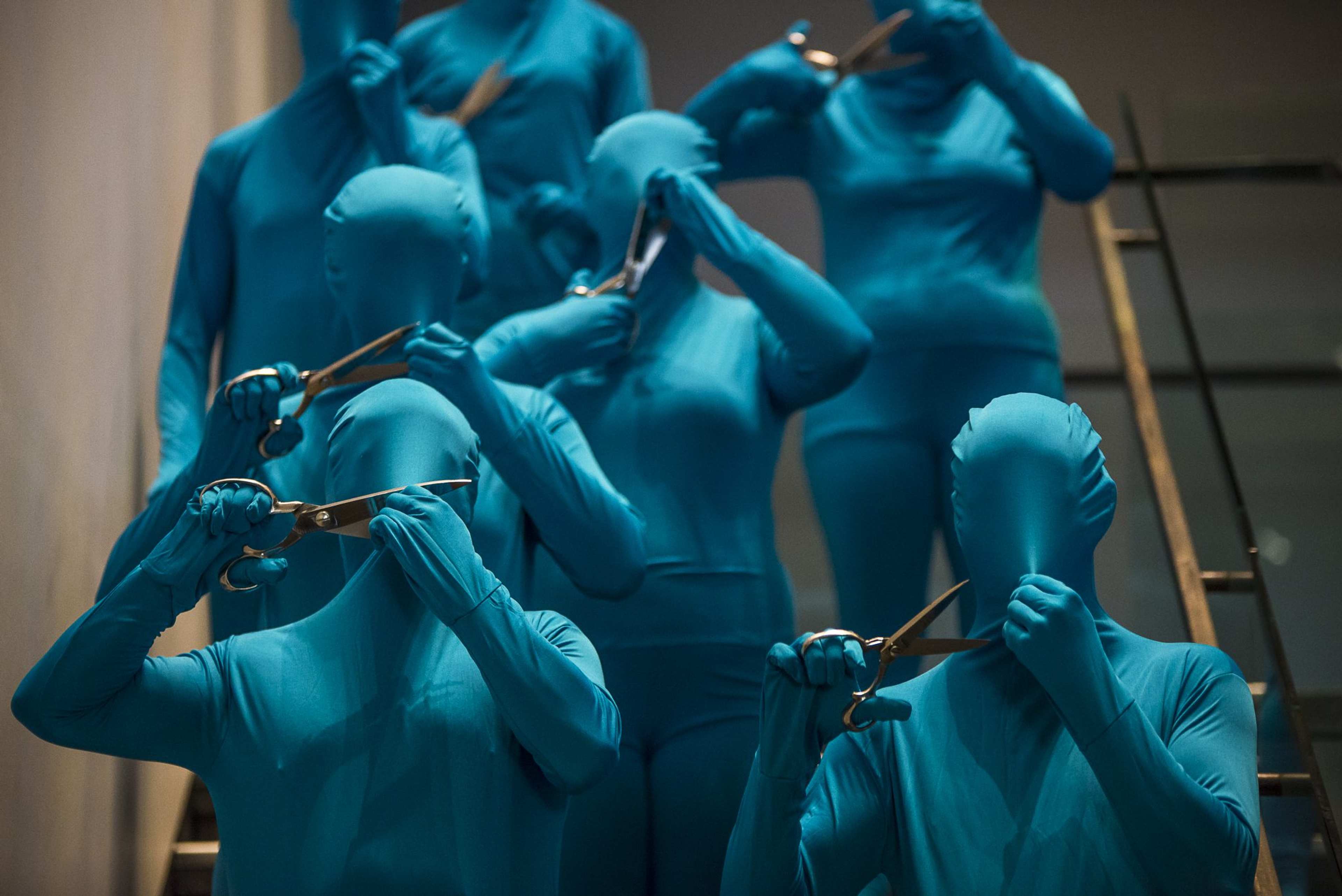 Eight performers in blue body suits use scissors to cut a hole in the fabric