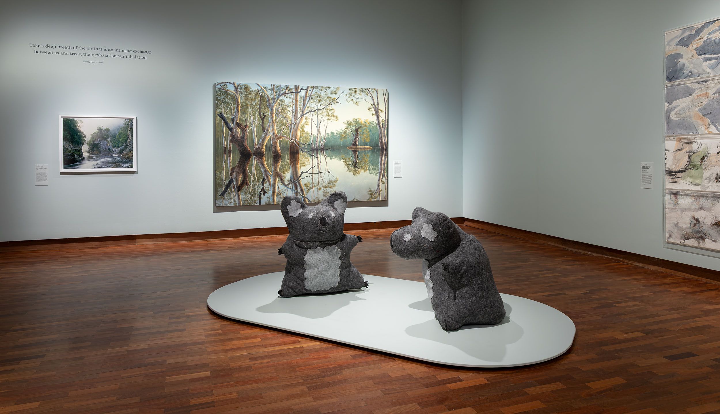 Two large plush koala sculptures on a white platform are displayed in an art gallery with wooden floors, surrounded by landscape paintings on light blue walls. Soft lighting highlights the artworks.