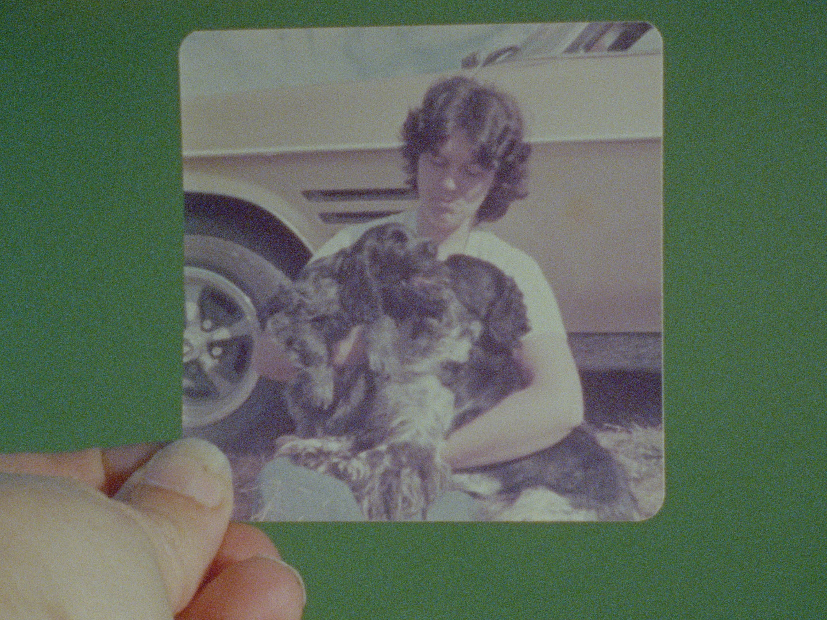 A hand holds a photo of a person with short brown hair sitting outdoors, holding two large black and white dogs in their lap. A cream-colored car is parked in the background.