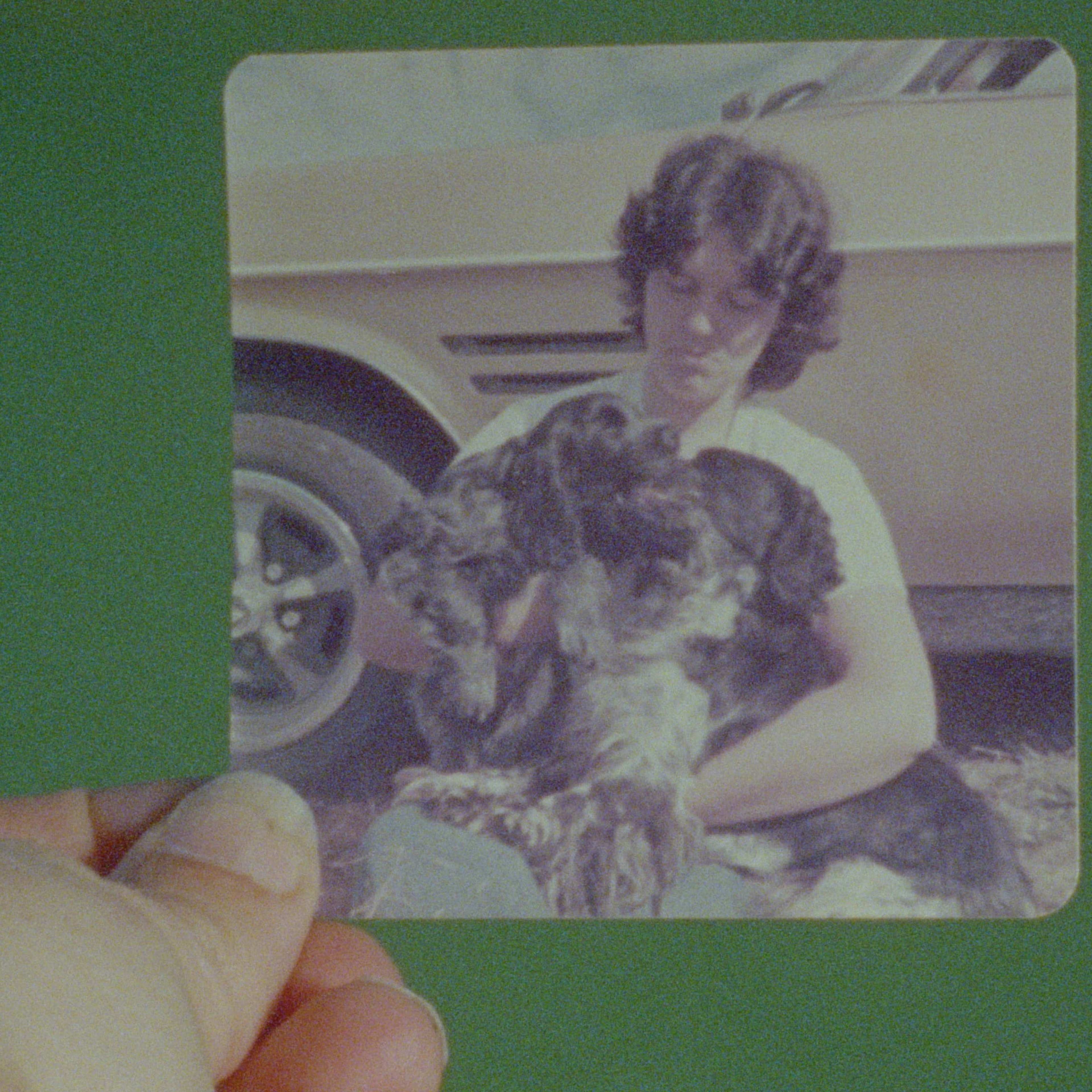 A hand holds a photo of a person with short brown hair sitting outdoors, holding two large black and white dogs in their lap. A cream-colored car is parked in the background.