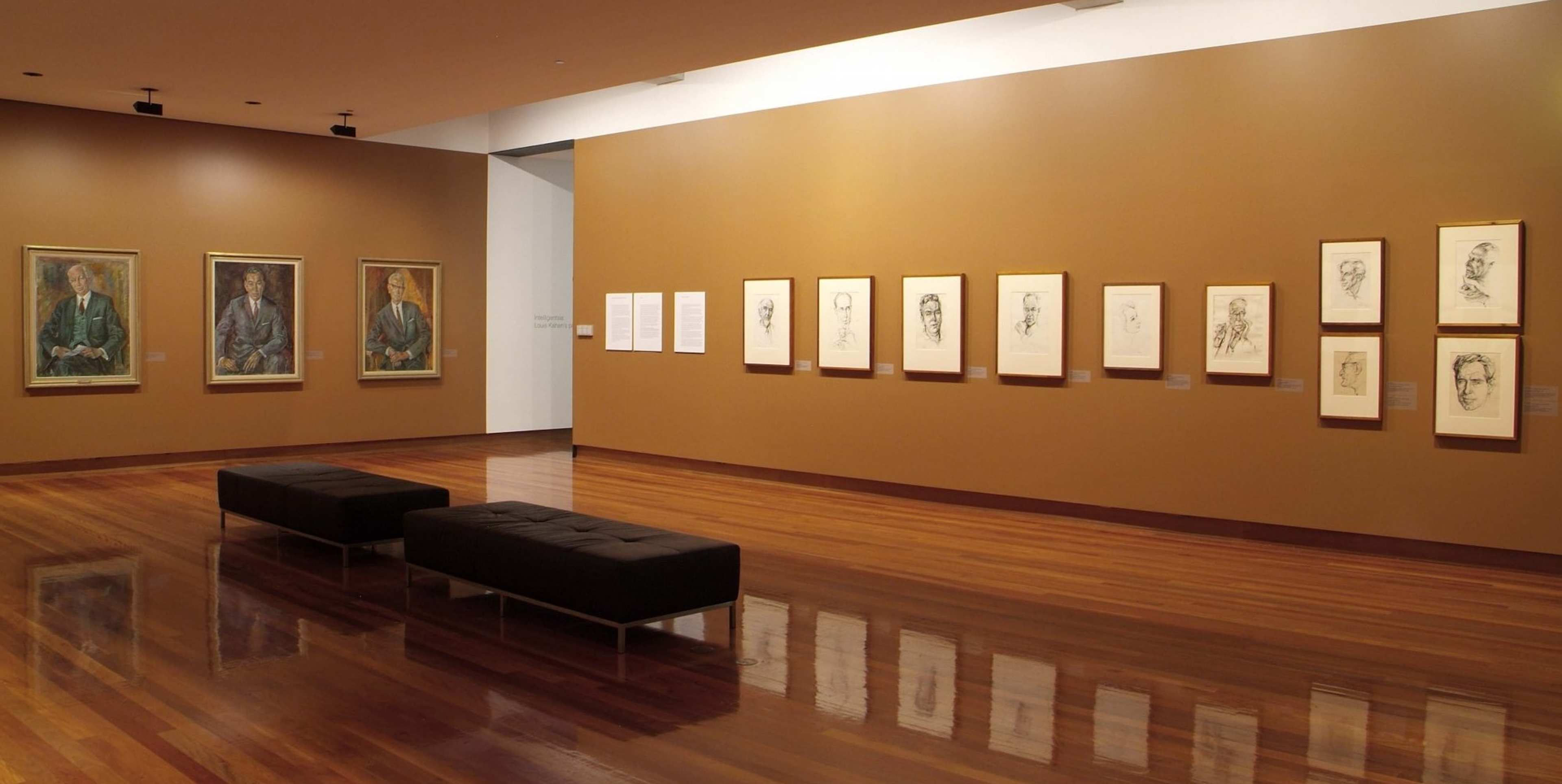 A photograph of a gallery space with walls painted mustard yellow and three colour portrait paintings on one wall and a row after black and white drawn portraits on the other side.