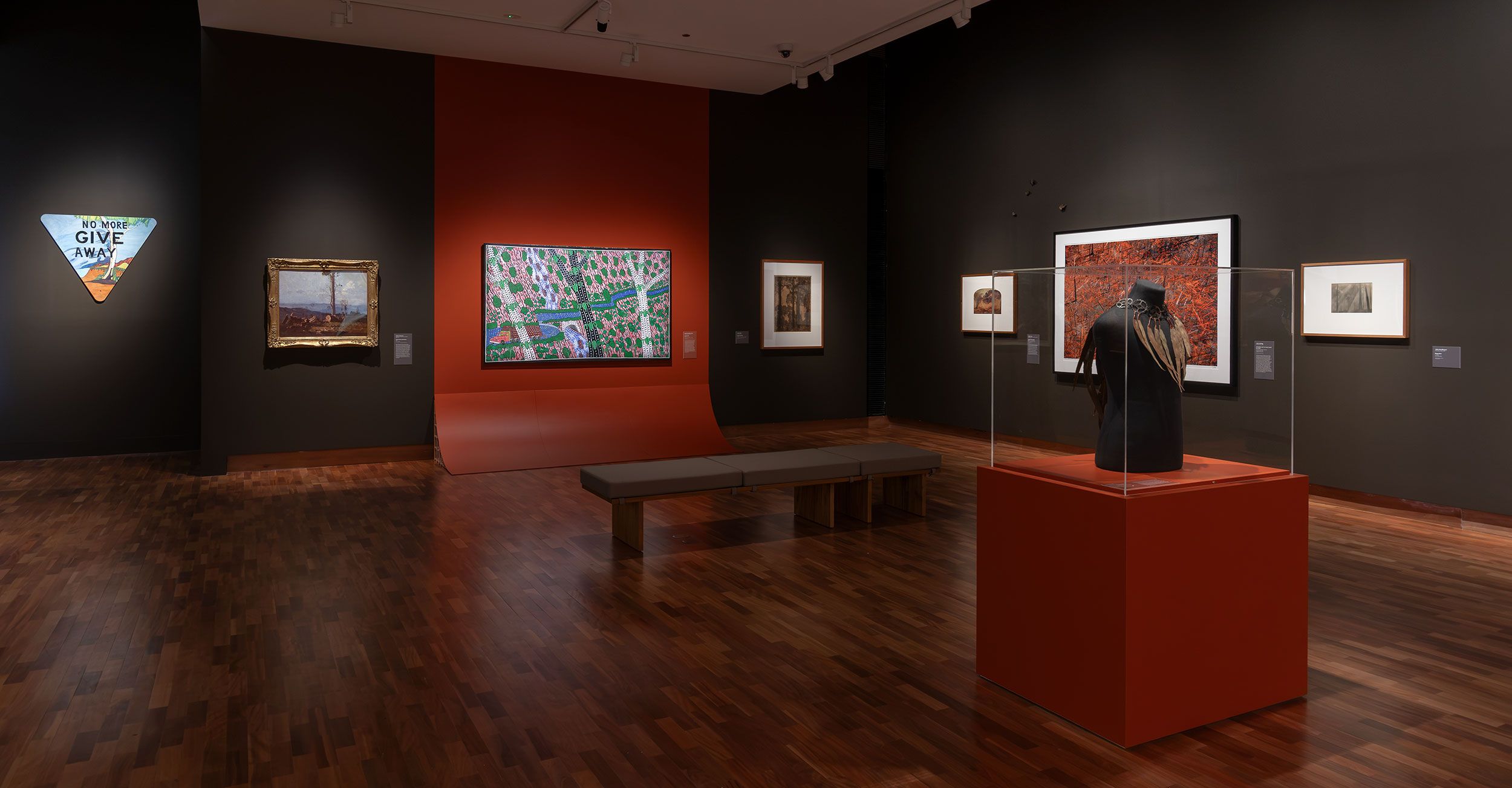 A modern art gallery with wooden floors, dark walls, and various artworks displayed, including paintings, a triangular sign, and a black garment in a glass case on an orange pedestal. A bench sits in the center of the room.