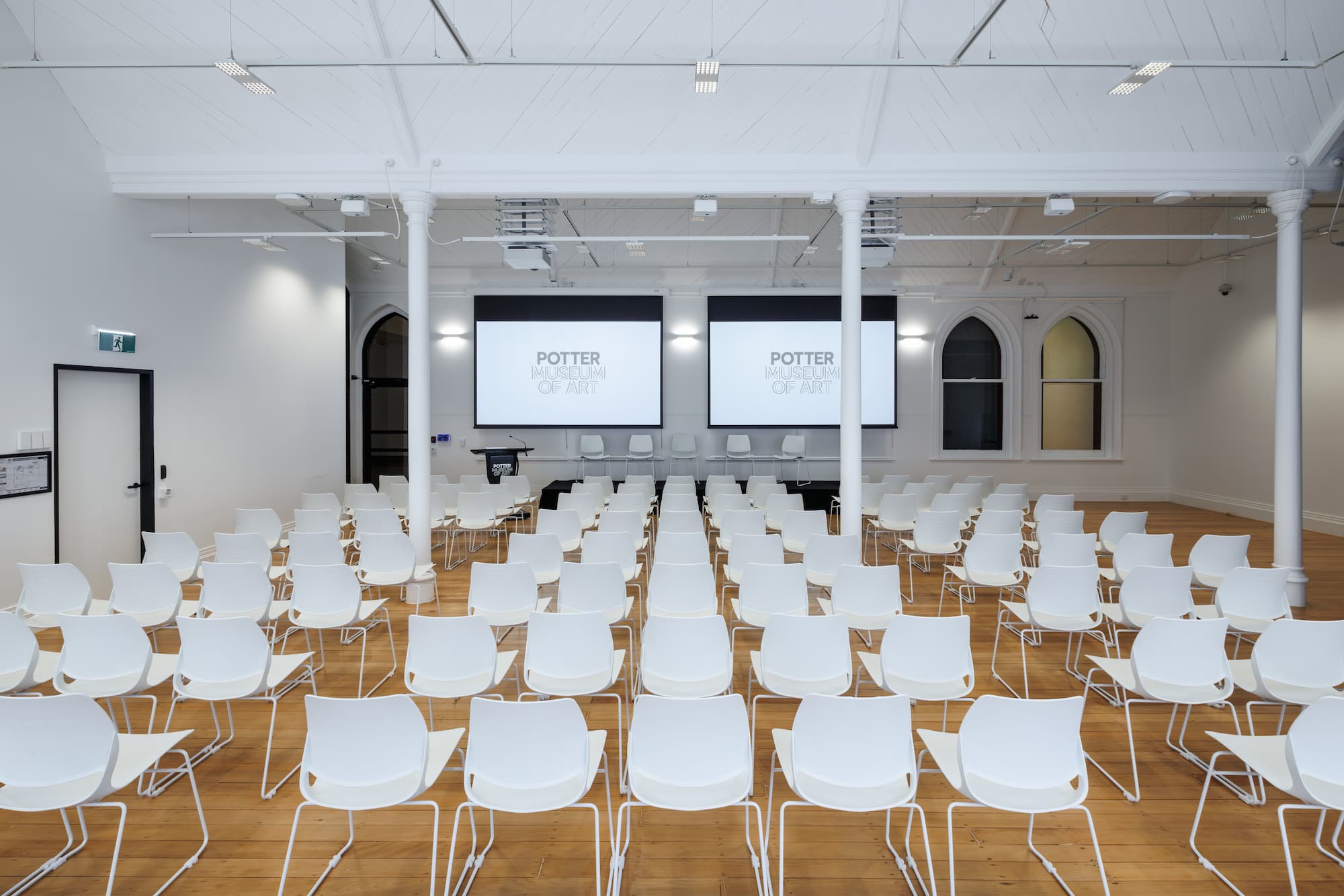 A wide room with lines of white chairs facing two screens at the front of the room with the Potter Museum of art logo