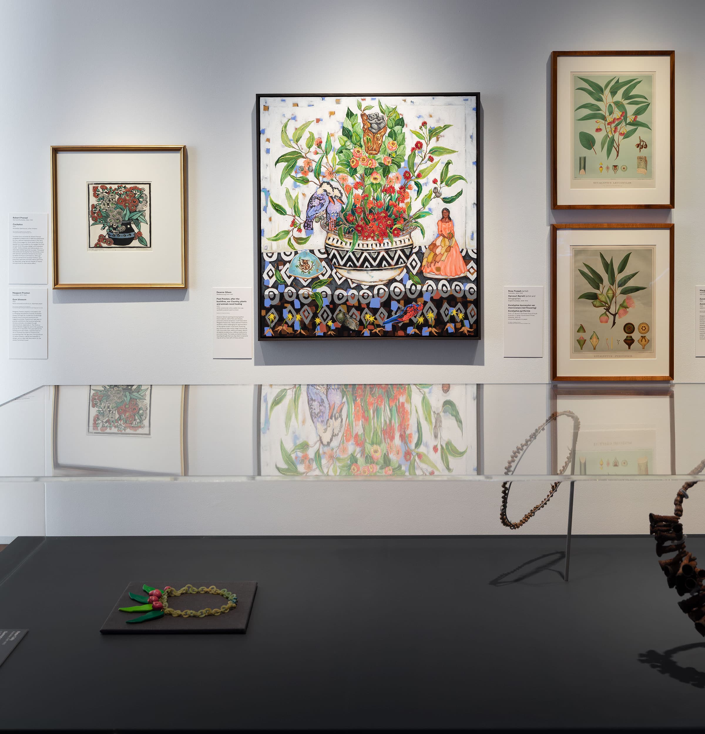 Framed botanical-themed artworks hang on a white wall above a display case containing jewelry pieces, including a necklace and brooch, all reflected on the glass surface.
