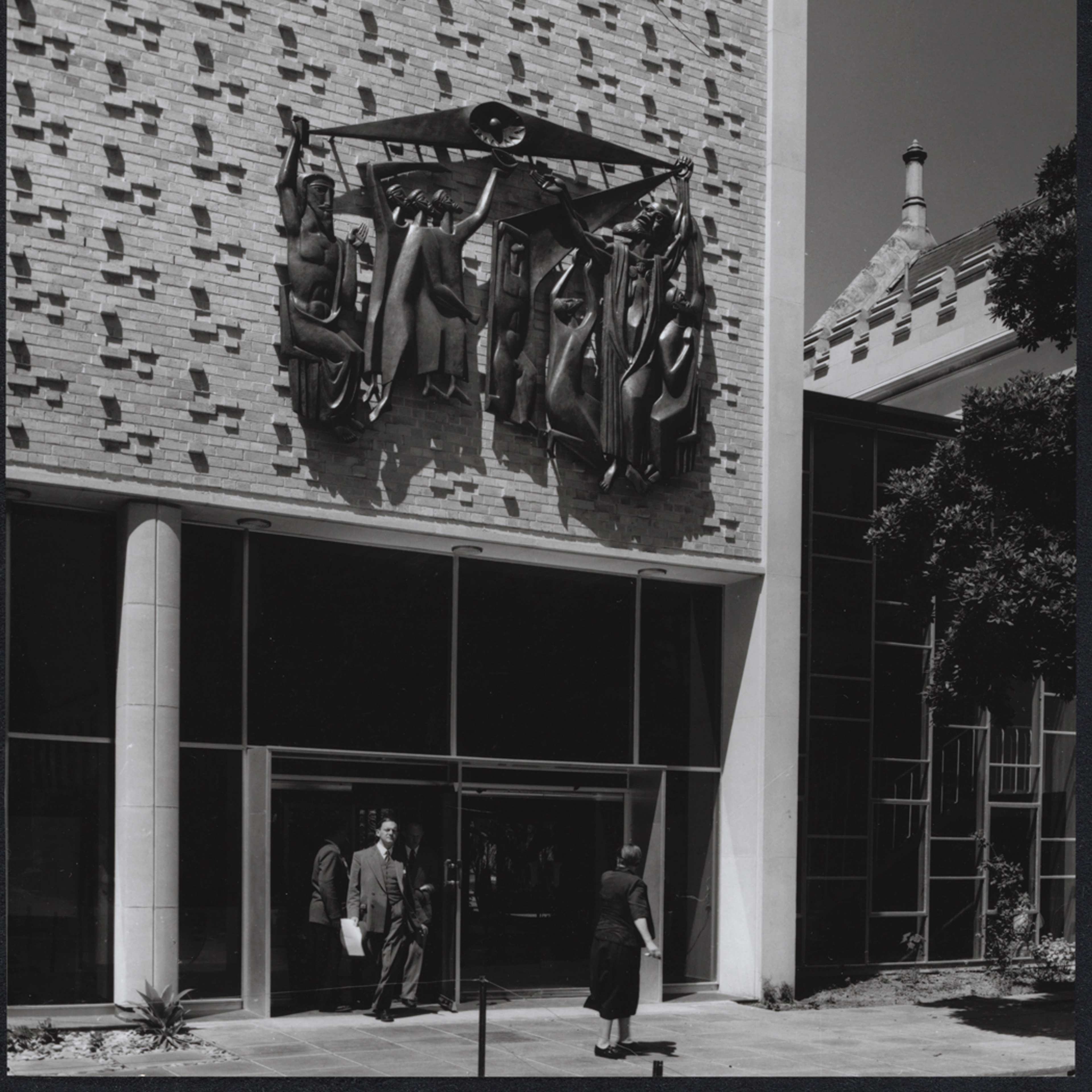A black and white photograph of an entrance to Wilson Hall at the University of Melbourne – a modernist-style building with glass doors and a sculptural panno above the entrance