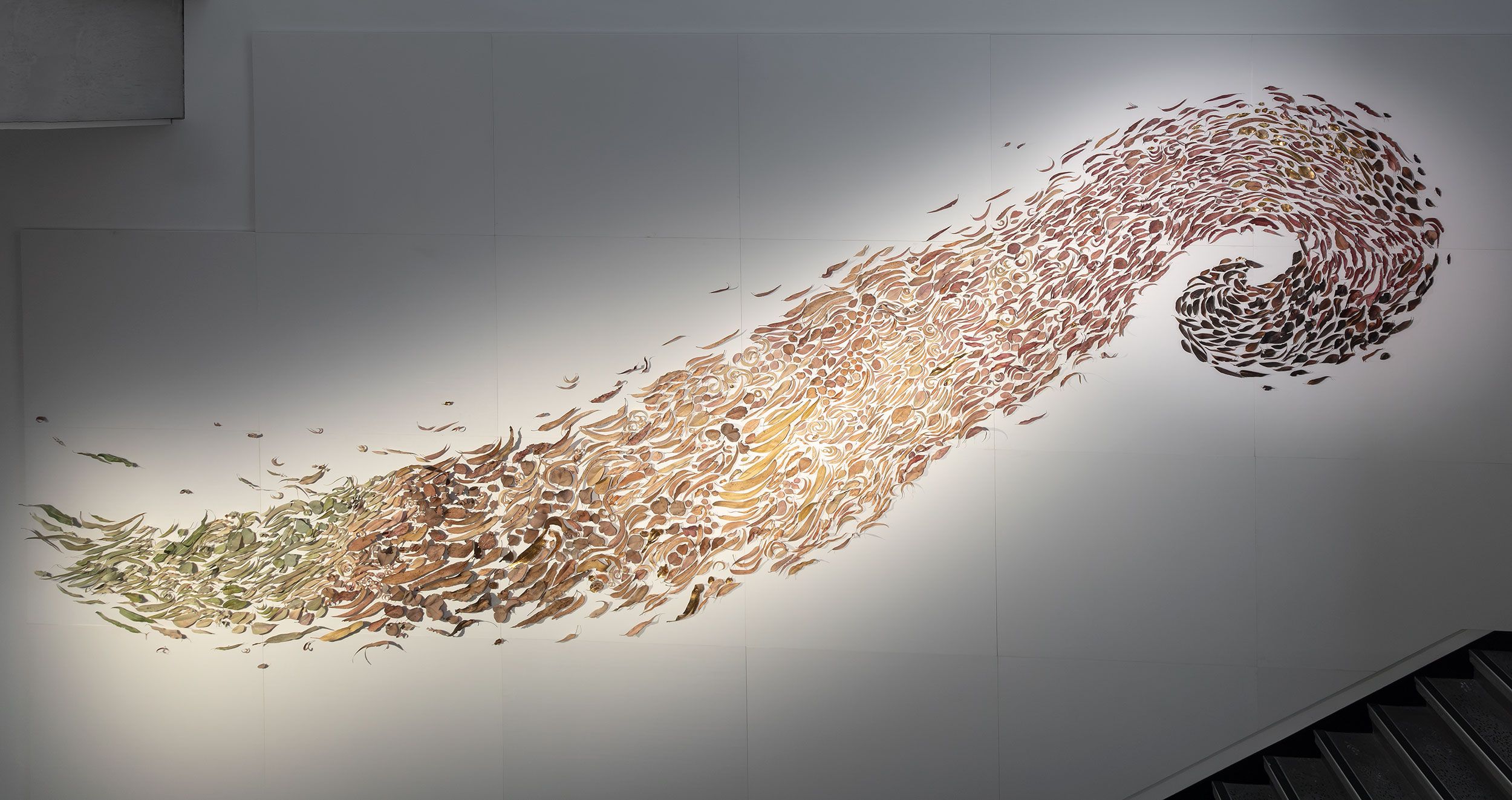 A large wall art installation shows hundreds of leaf-shaped pieces arranged in a swirling, wave-like pattern, transitioning in color from green to brown.