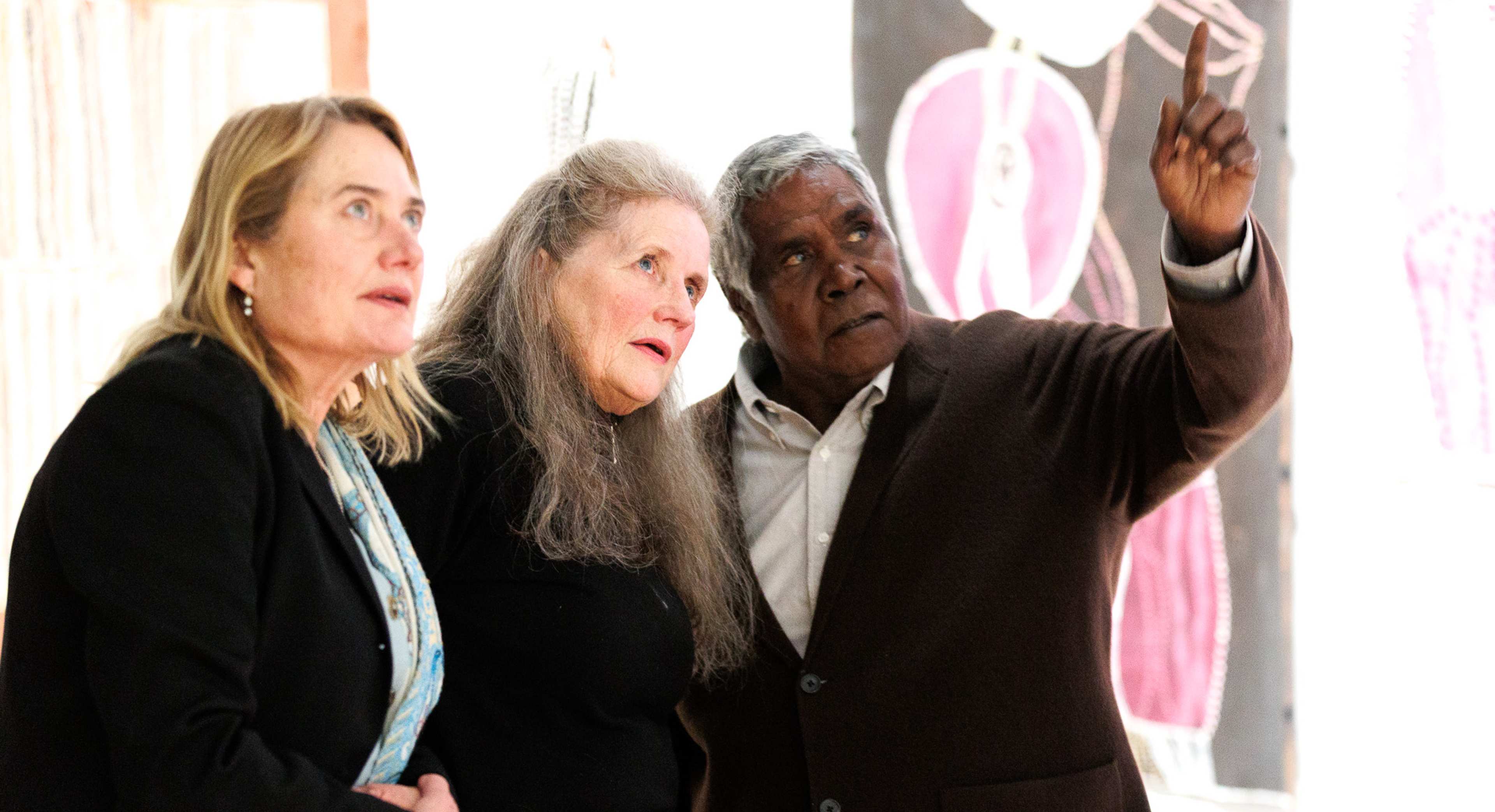 Three people stand closely together in an art gallery, looking intently in the same direction as a man on the right points upwards. Colourful abstract paintings are visible on the wall behind them.