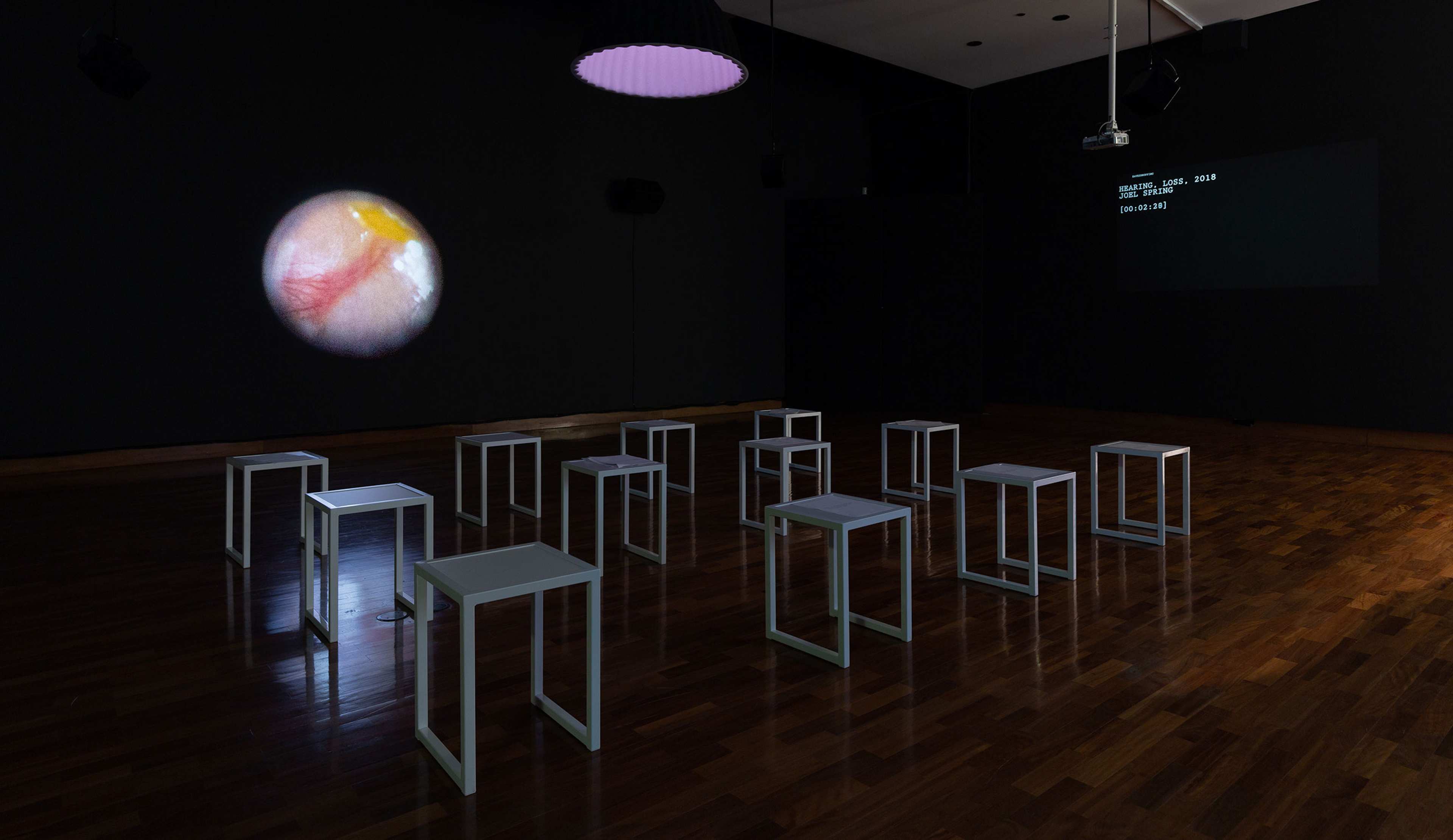 A darkened gallery space with white stools and two projected artworks, one displaying a text that  is hard to read and another an outline of a planet