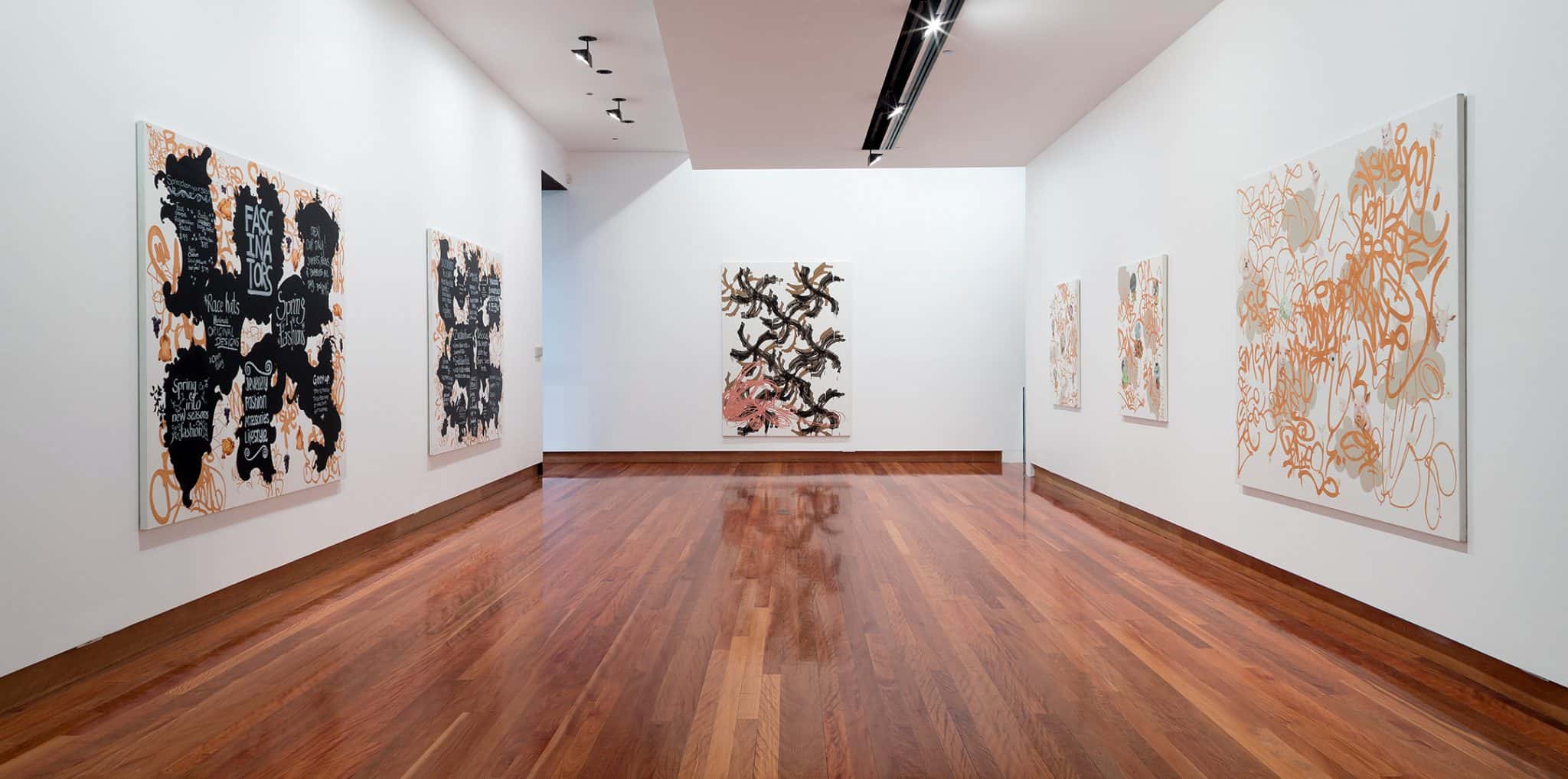 Large abstract canvases in black, brown and gold line up white walls of a gallery space