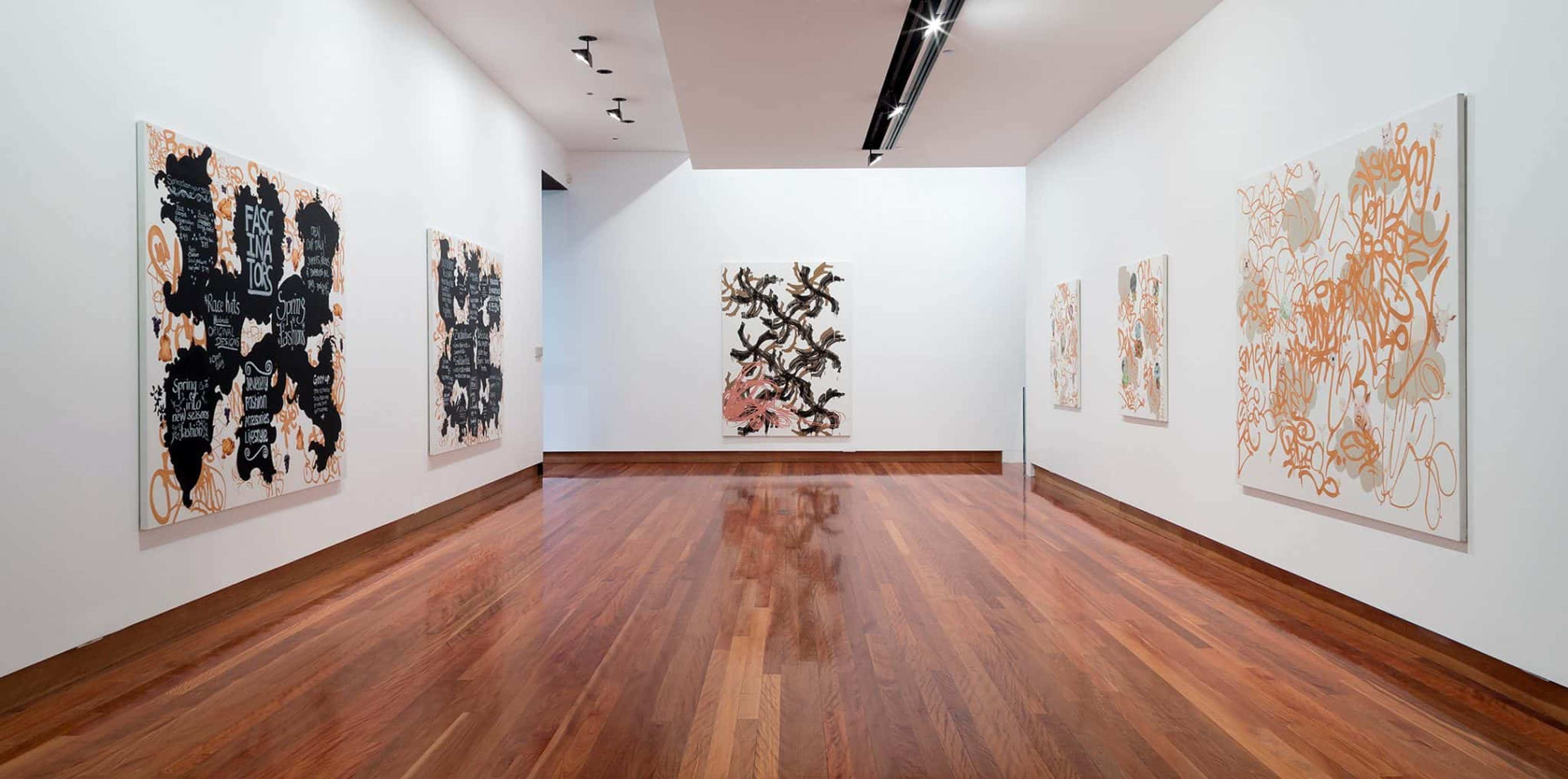 Large abstract canvases in black, brown and gold line up white walls of a gallery space