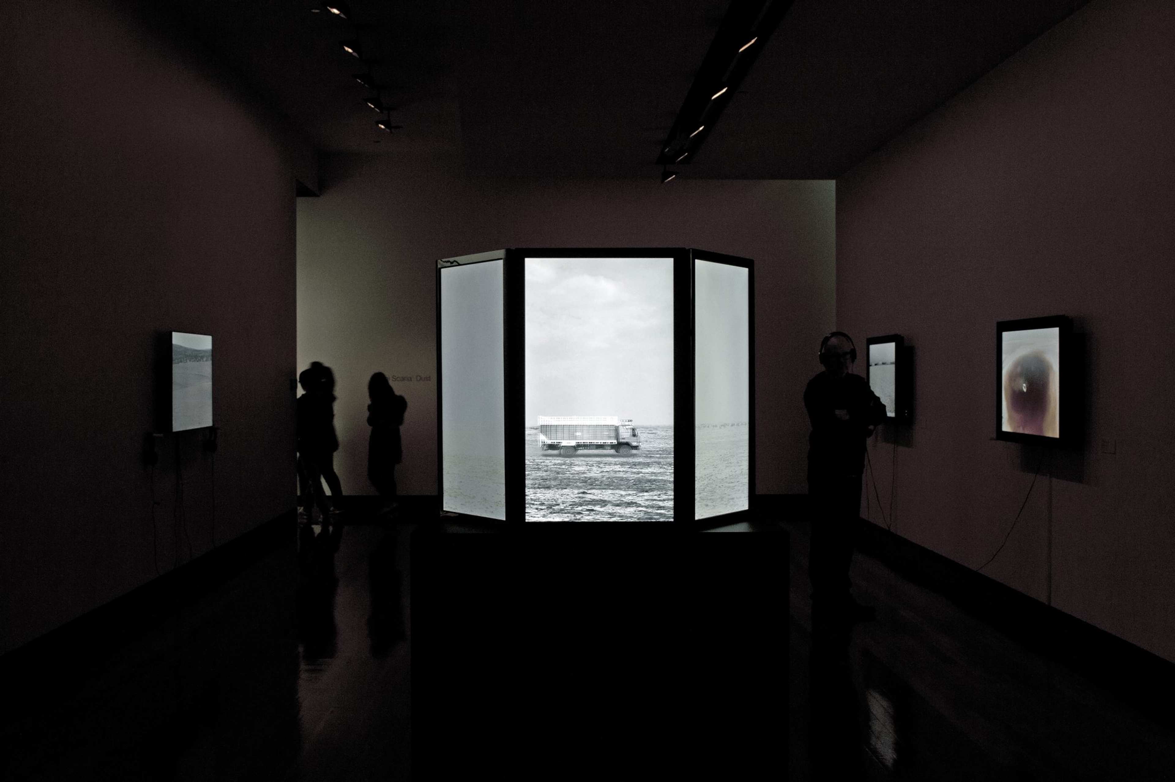 A photograph of a darkened gallery space with wall-mounted lightboxes