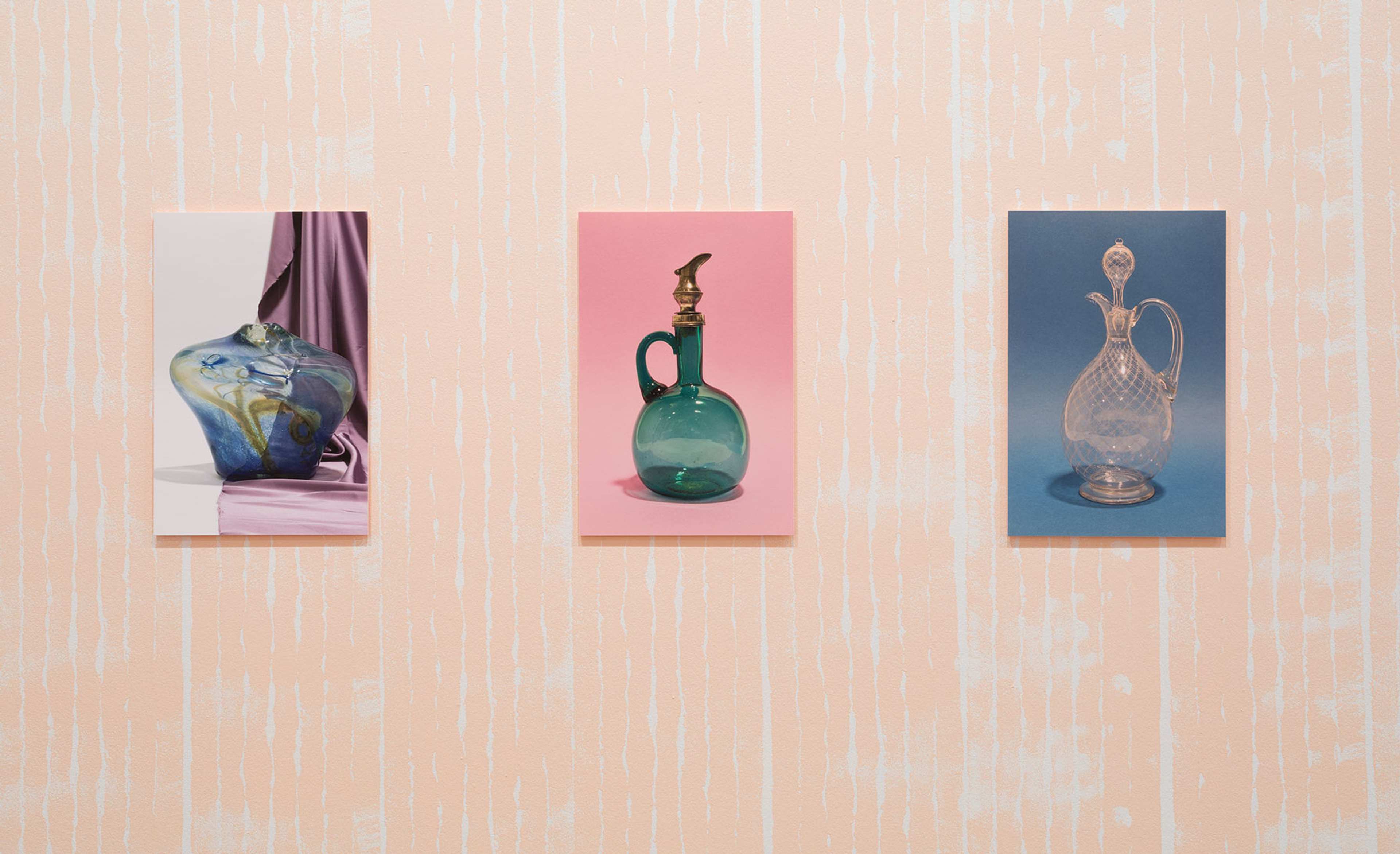 Three photographs of ornate glass jugs and a carafe hang in line on a pink wall
