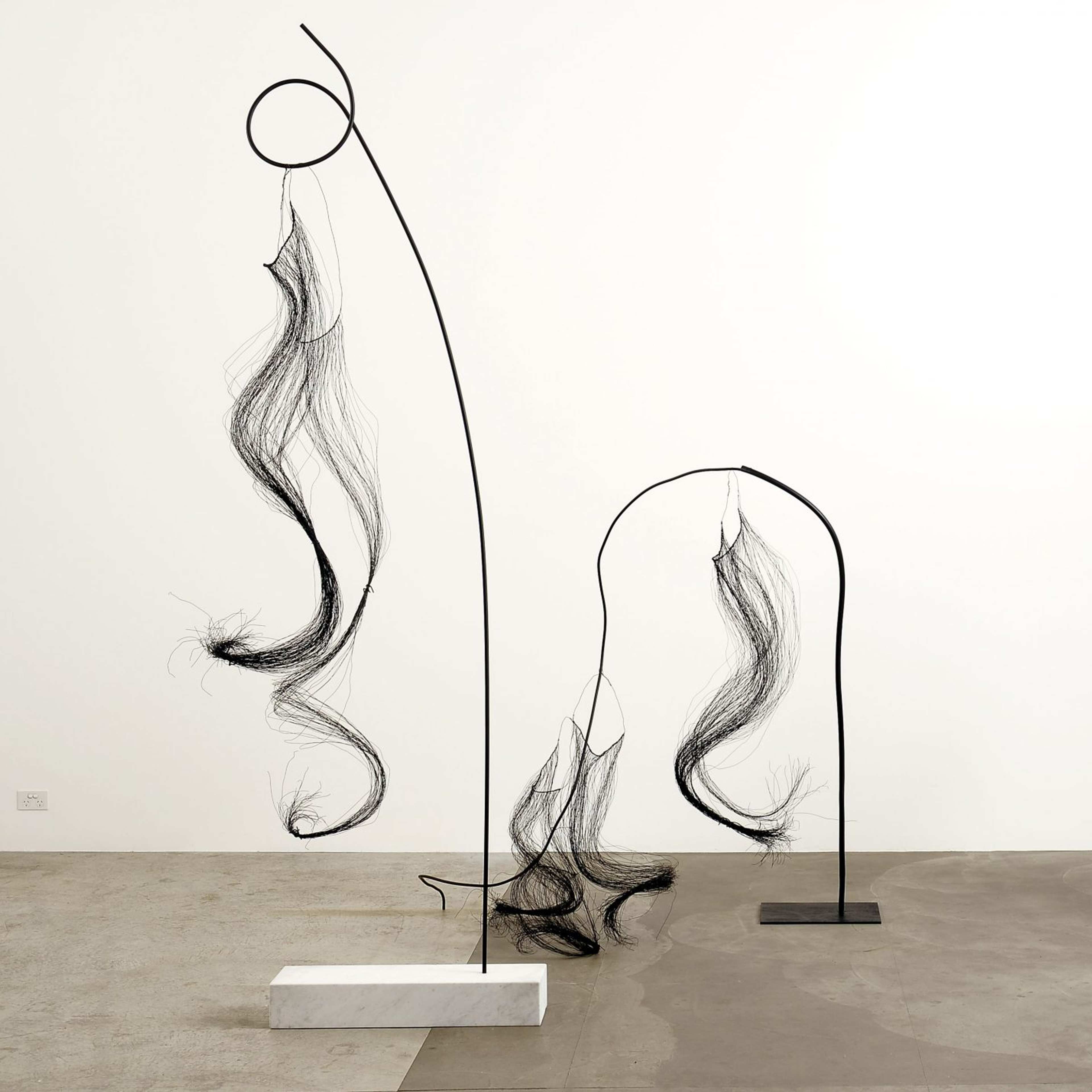 A photograph of three sculptural works consisting of vertical rods and strands of thin wires that look like curly hair