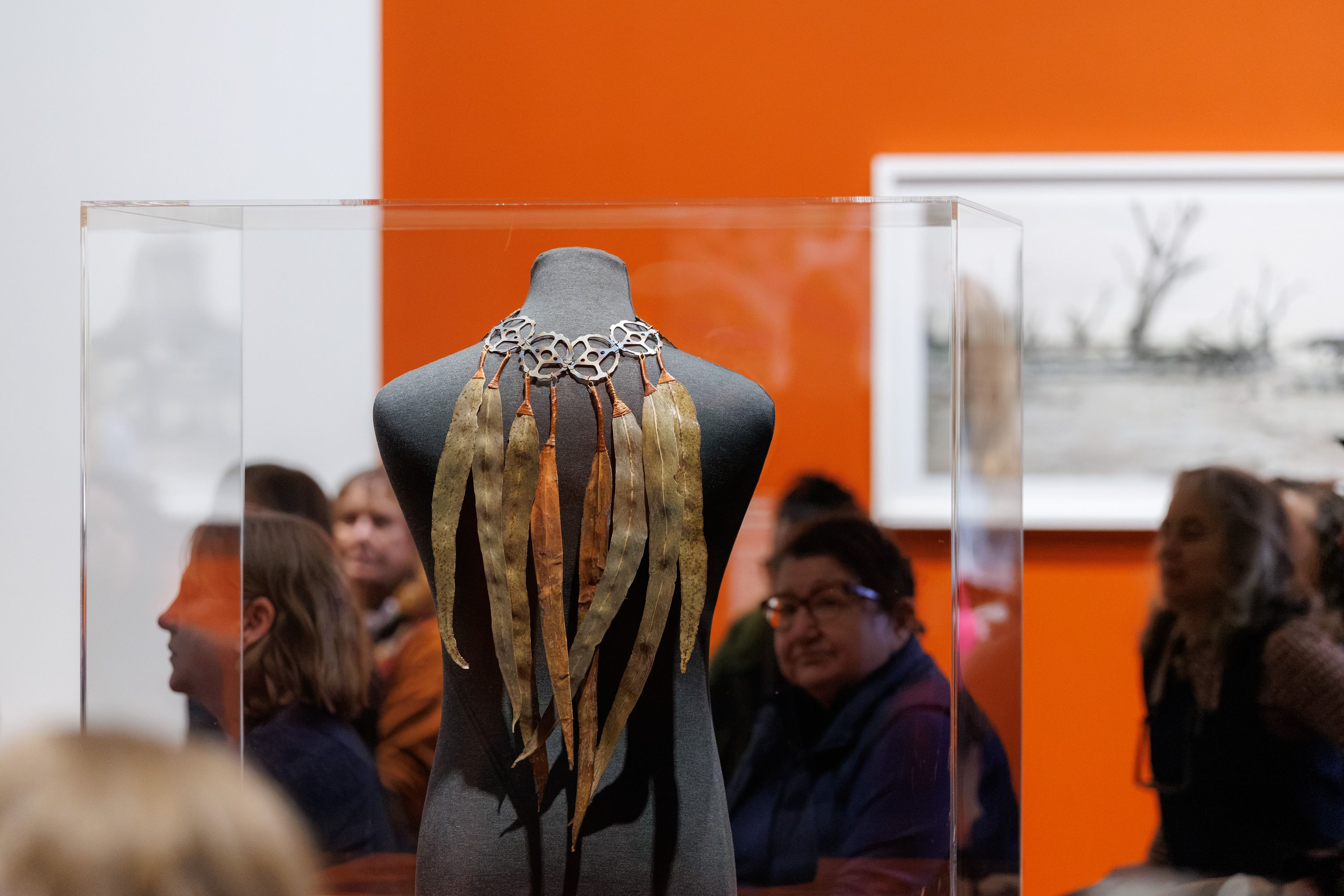 A gallery room with a work of gum leaves hooked onto sewing machine cogs, attached to form an accessory, draping over the back of a mannequin body in a clear casing.