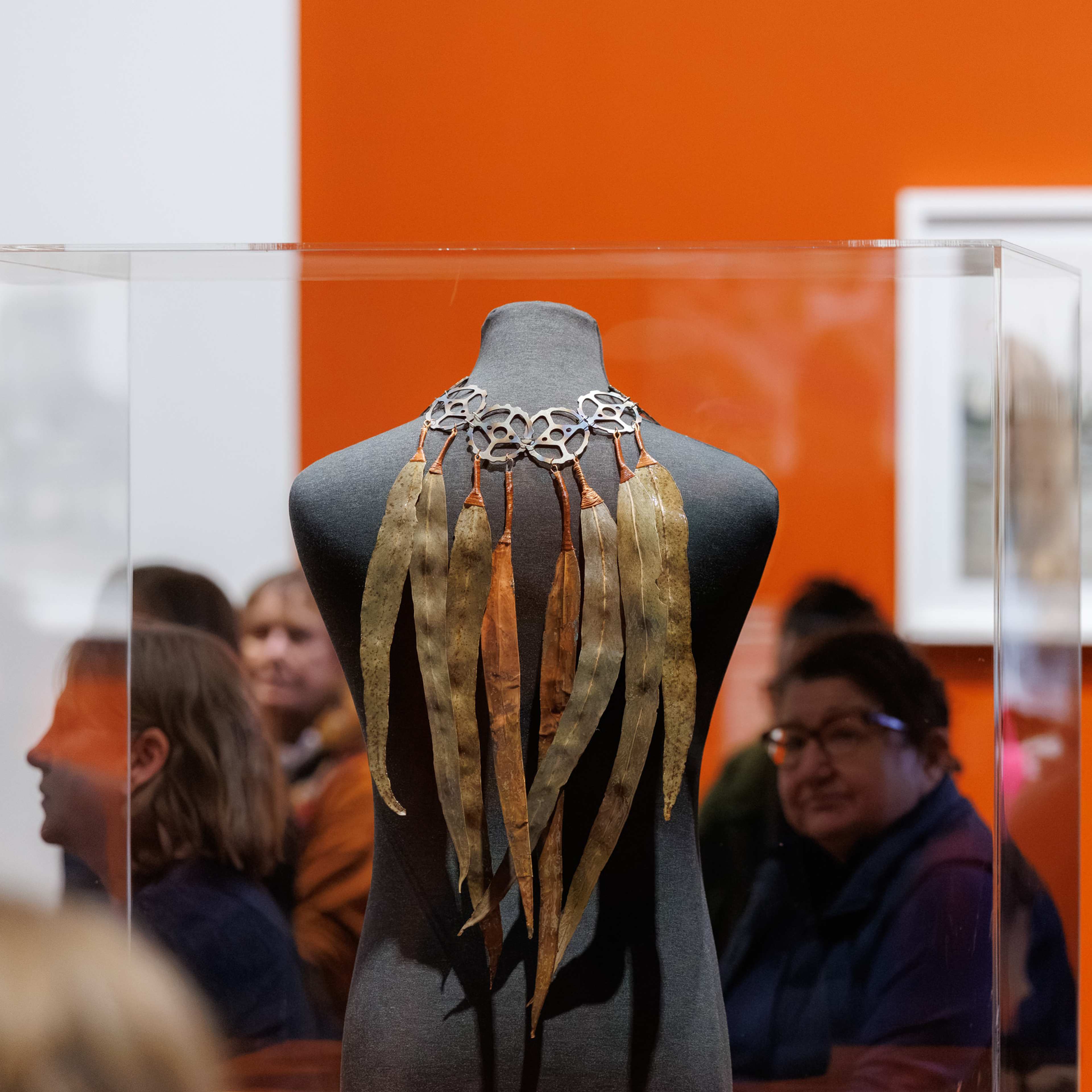 A gallery room with a work of gum leaves hooked onto sewing machine cogs, attached to form an accessory, draping over the back of a mannequin body in a clear casing.