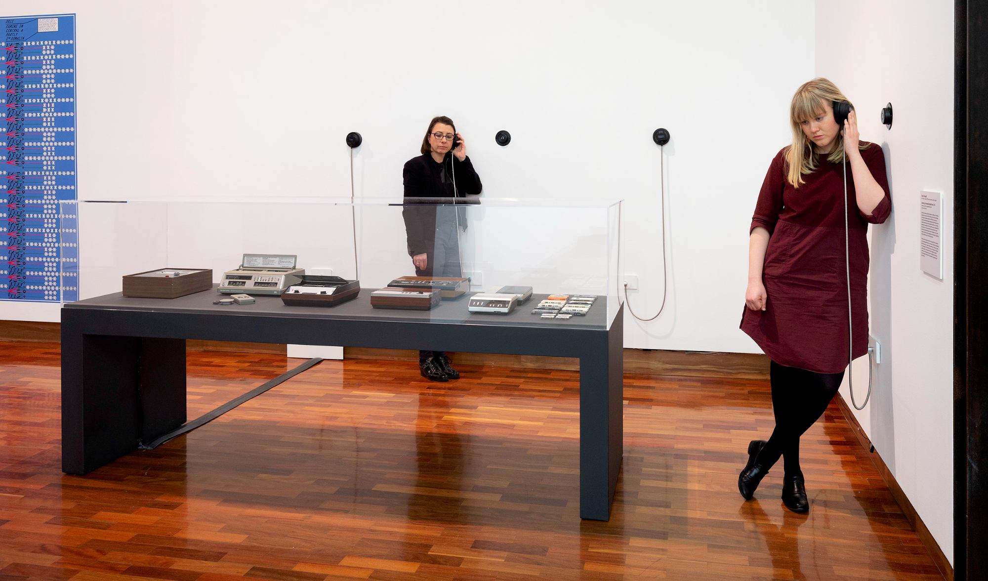 Two women listen to headphones attached to a wall in a gallery, old recording devices in a plinth