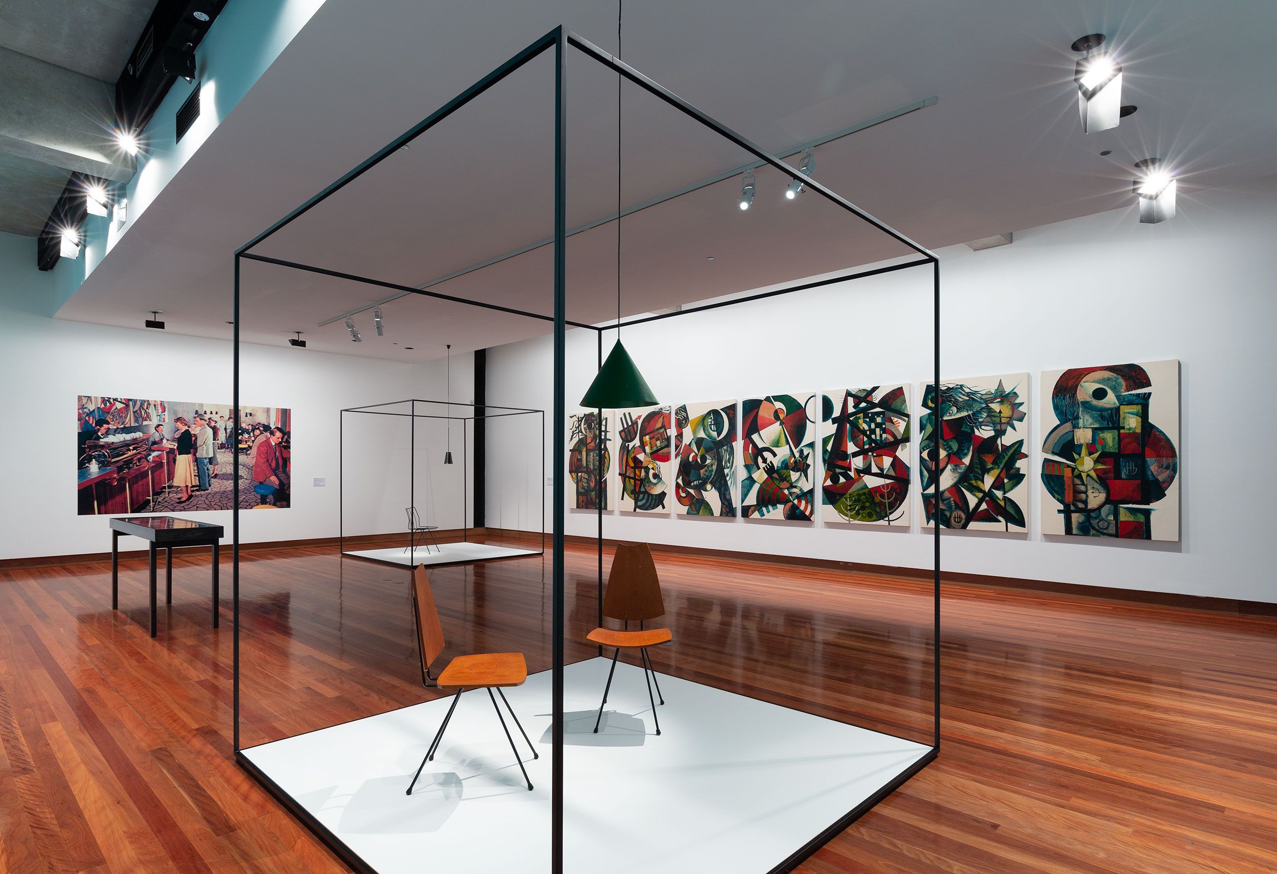 A wide shot of a gallery with mid century chairs and tables laid out in a gallery with paintings on walls