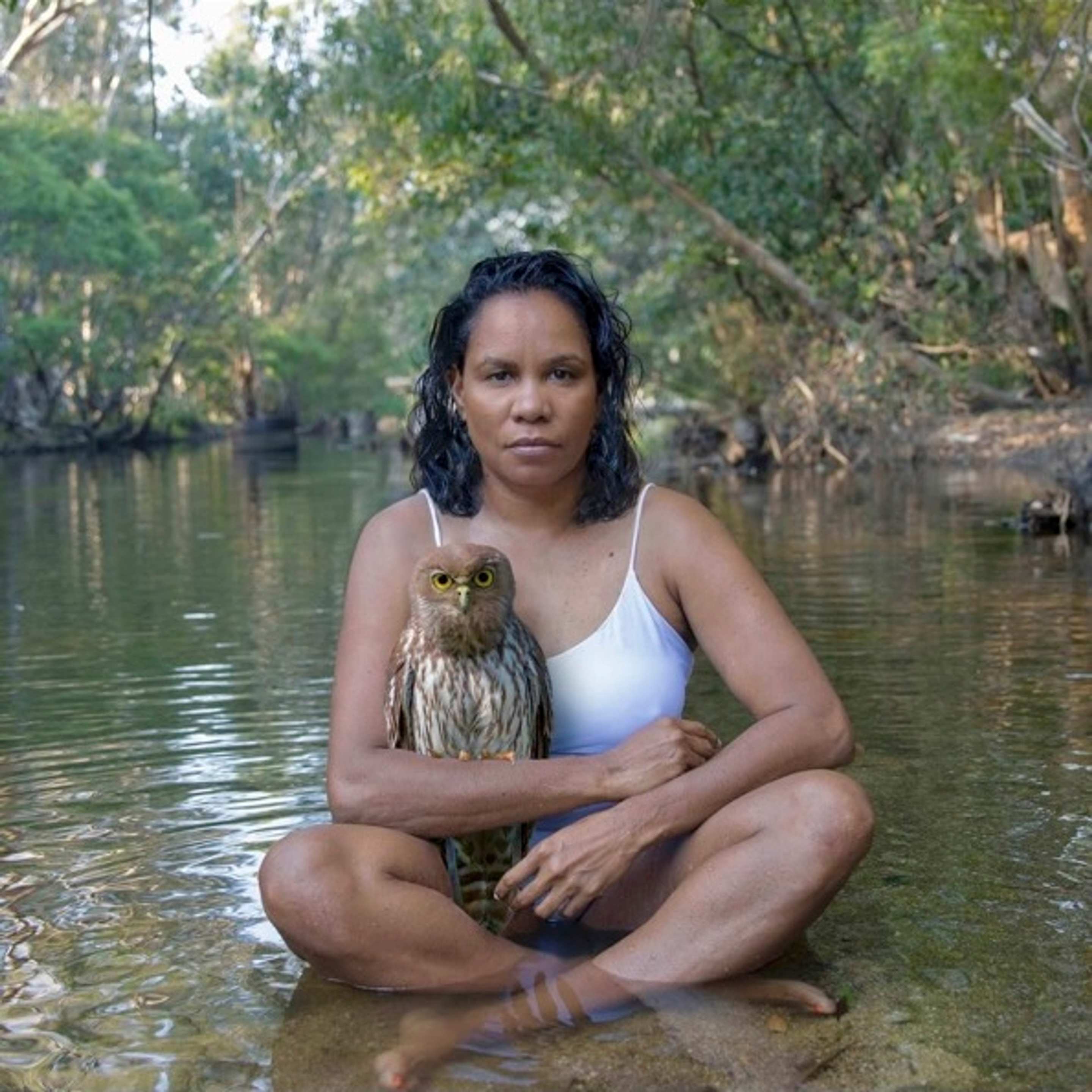 A person sits in a river in a bushy landscape in a white singlet with a brown owl sitting on their arm.