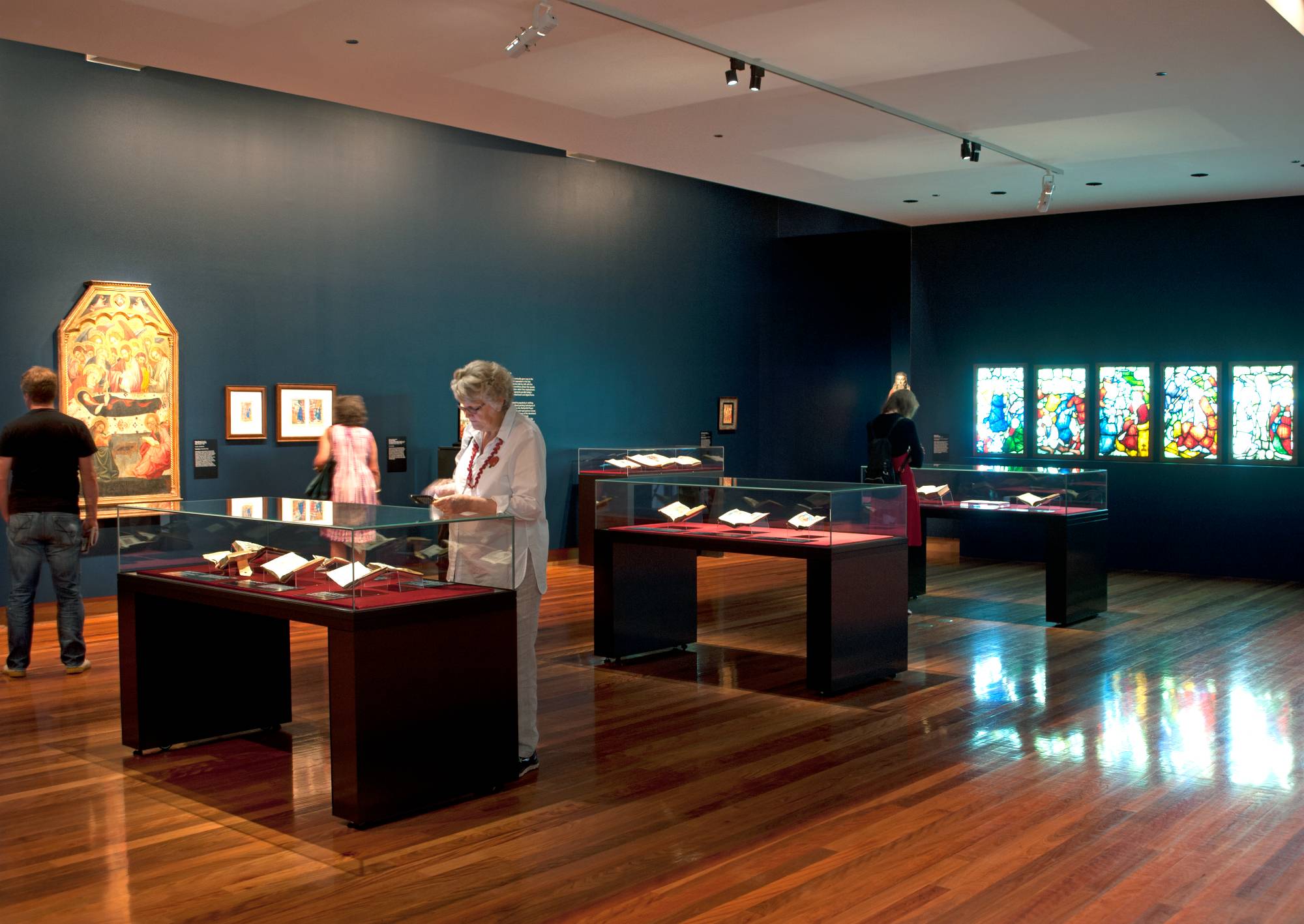 A darkened gallery with a row of vitrines, a medieval painting on one of the black-painted walls and a row os stained glass panels on the other