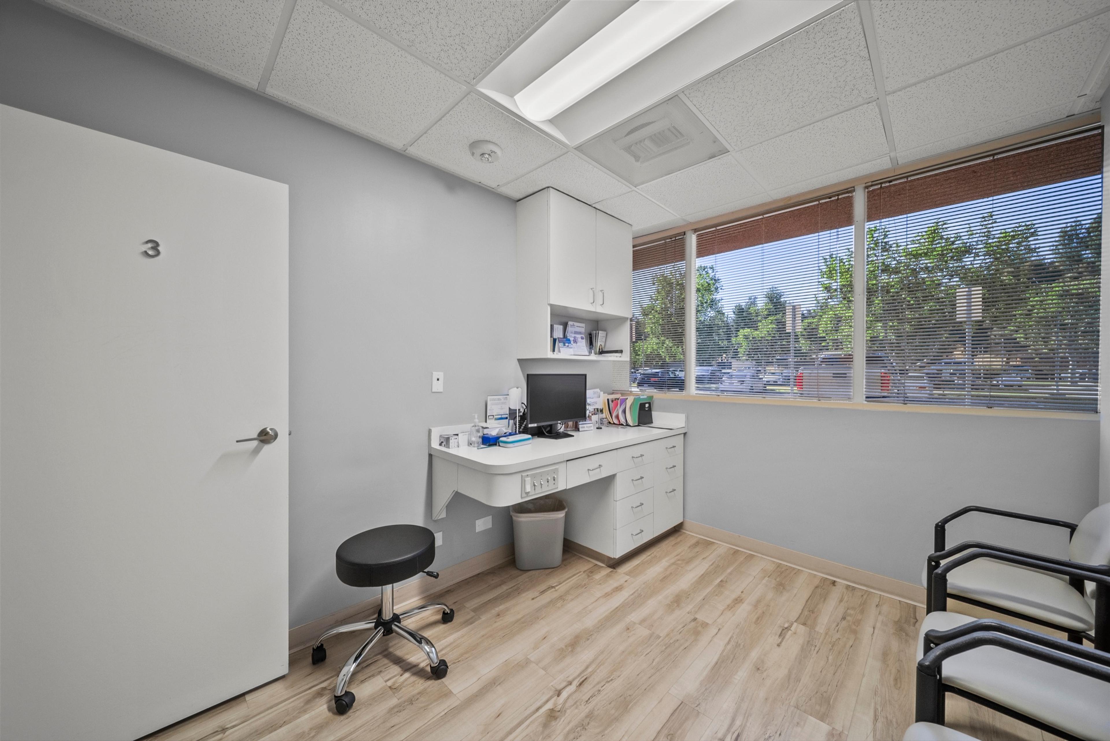 Exam room 3, with a built-in workstation and computer, a clinician’s stool, and a large window overlooking trees.