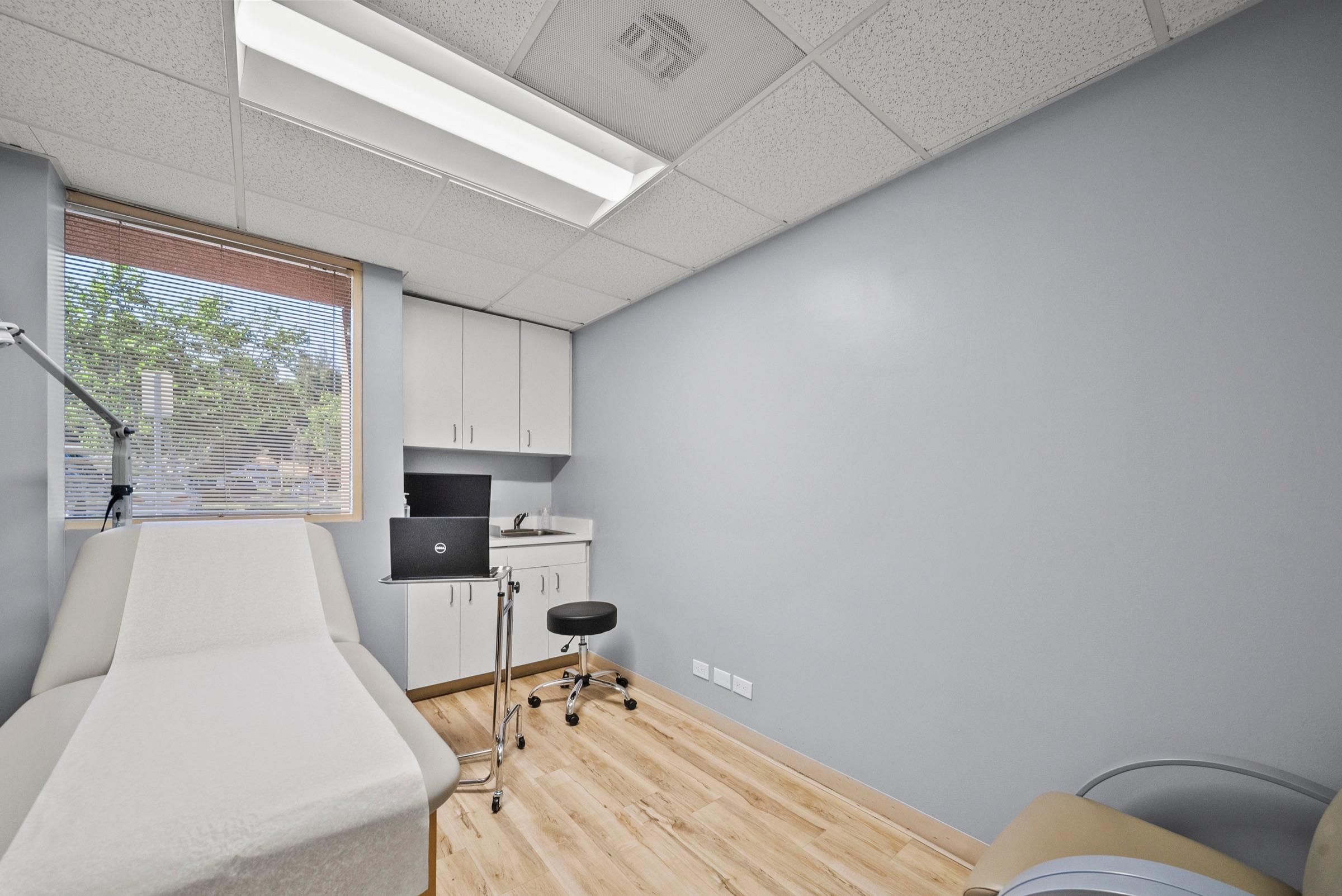 A neutral exam room with a paper-covered exam table beside a window, a laptop on a rolling stand, and a clinician’s stool.