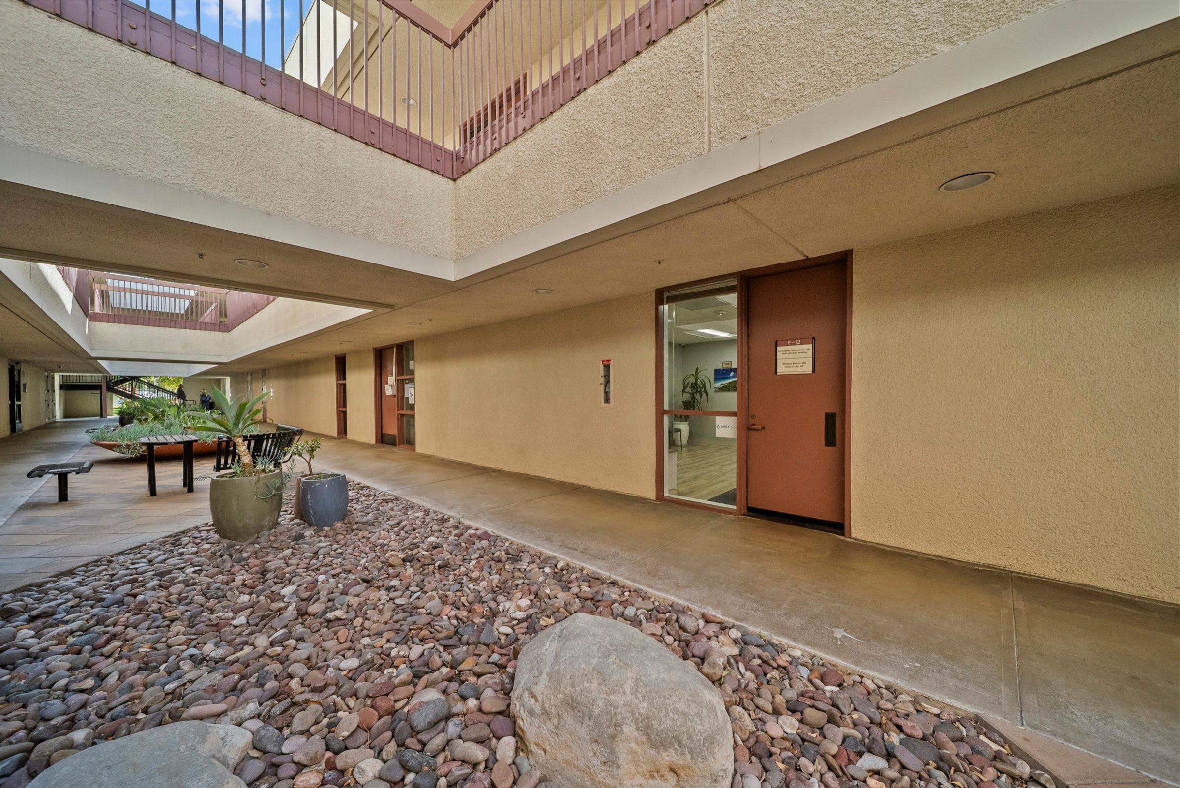 The outdoor courtyard entrance to the practice, with river-rock landscaping, potted plants, and the glass office door marked E-12.