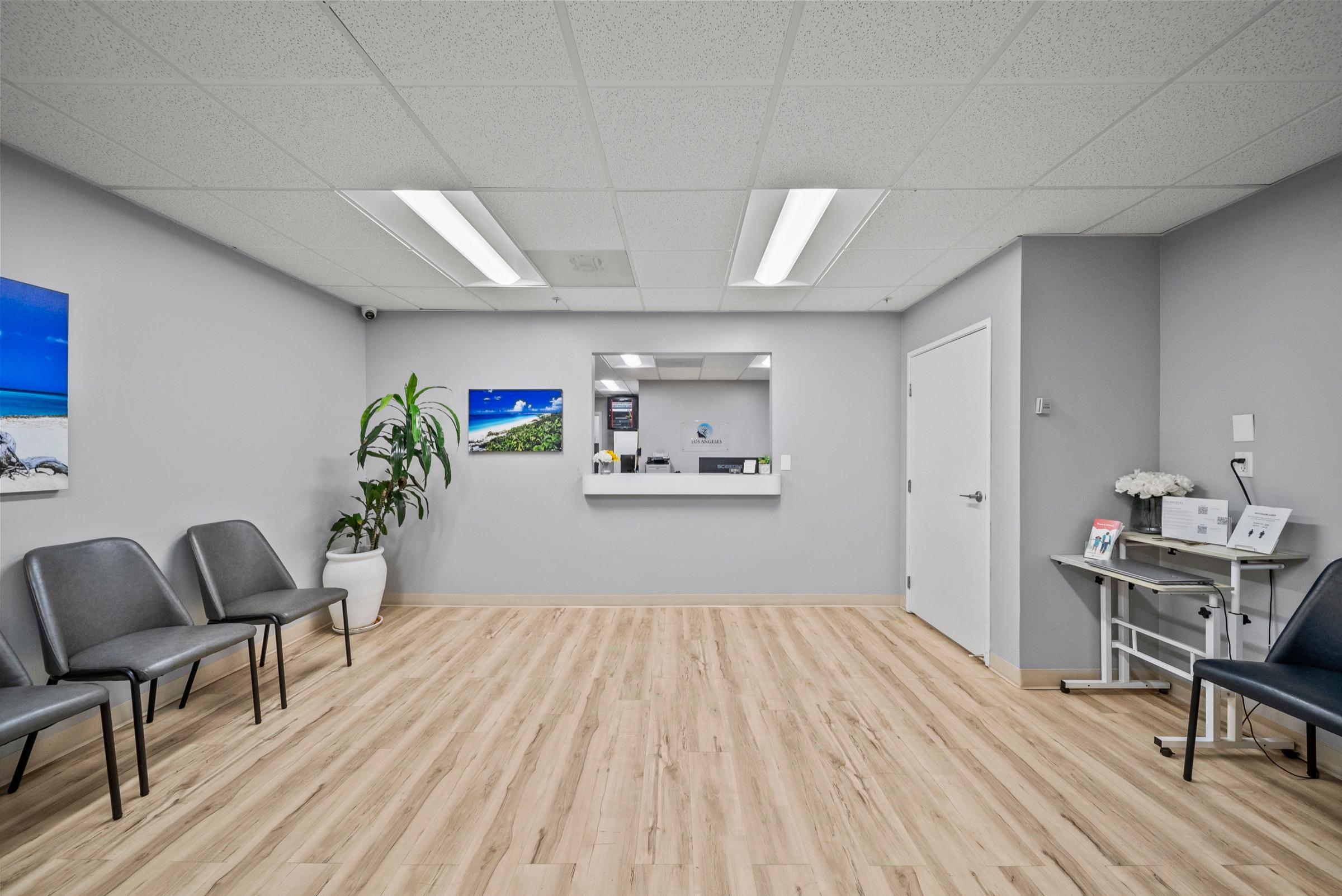 The Los Angeles Neurosciences reception and waiting area — a bright room with light wood floors, gray seating, framed coastal artwork, and a check-in window with the practice logo.