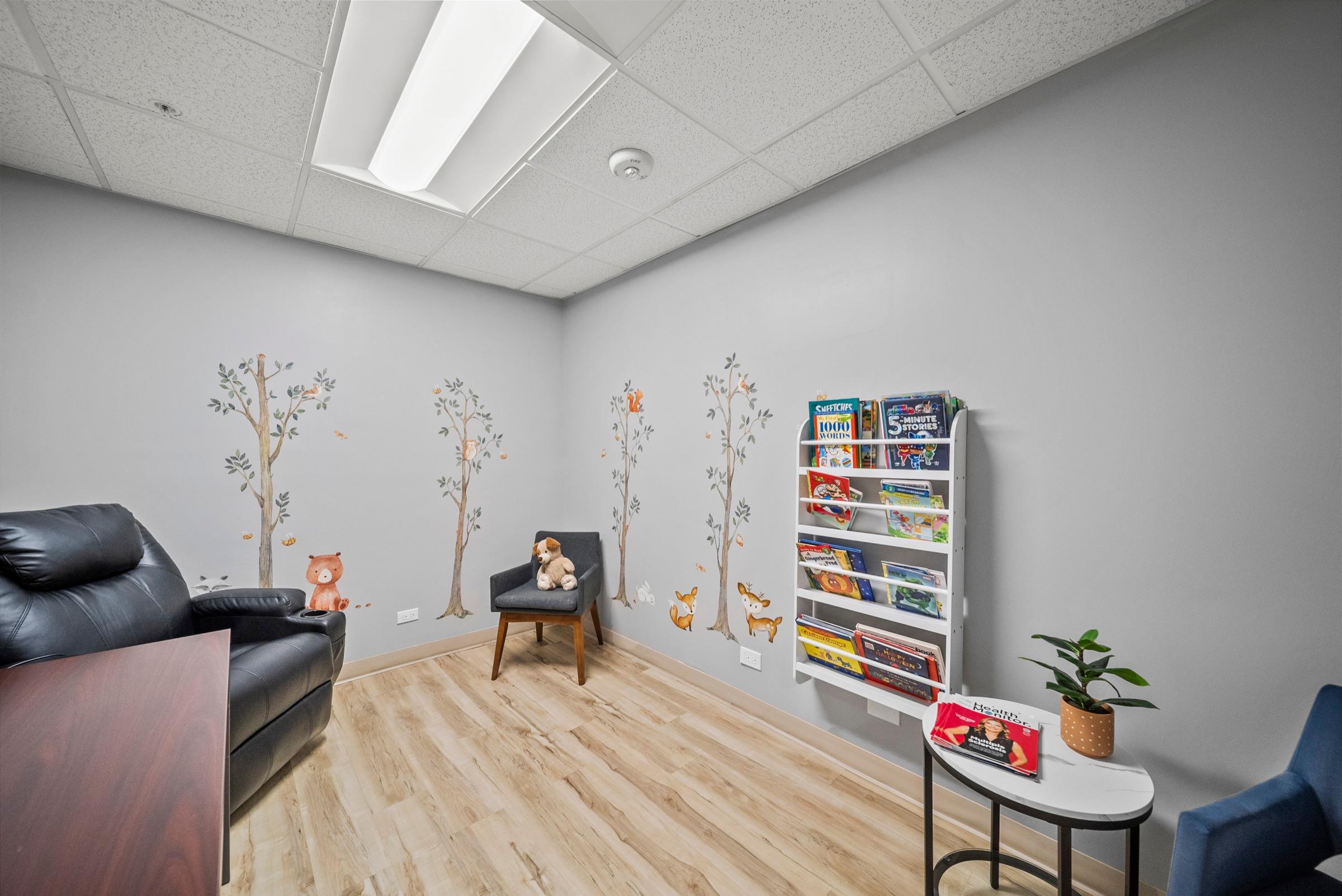 A child-friendly exam room with woodland tree-and-animal wall art, soft seating, and a rack of children’s books.