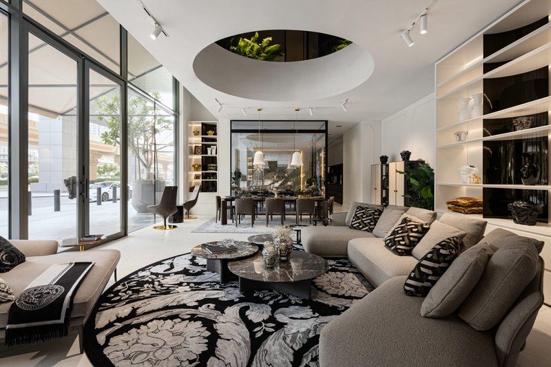 A sophisticated woman in a bright, modern Dubai apartment sitting on a designer sofa and browsing luxury furniture collections on a tablet, with marble elements, brass accessories, and a Dubai Marina view.