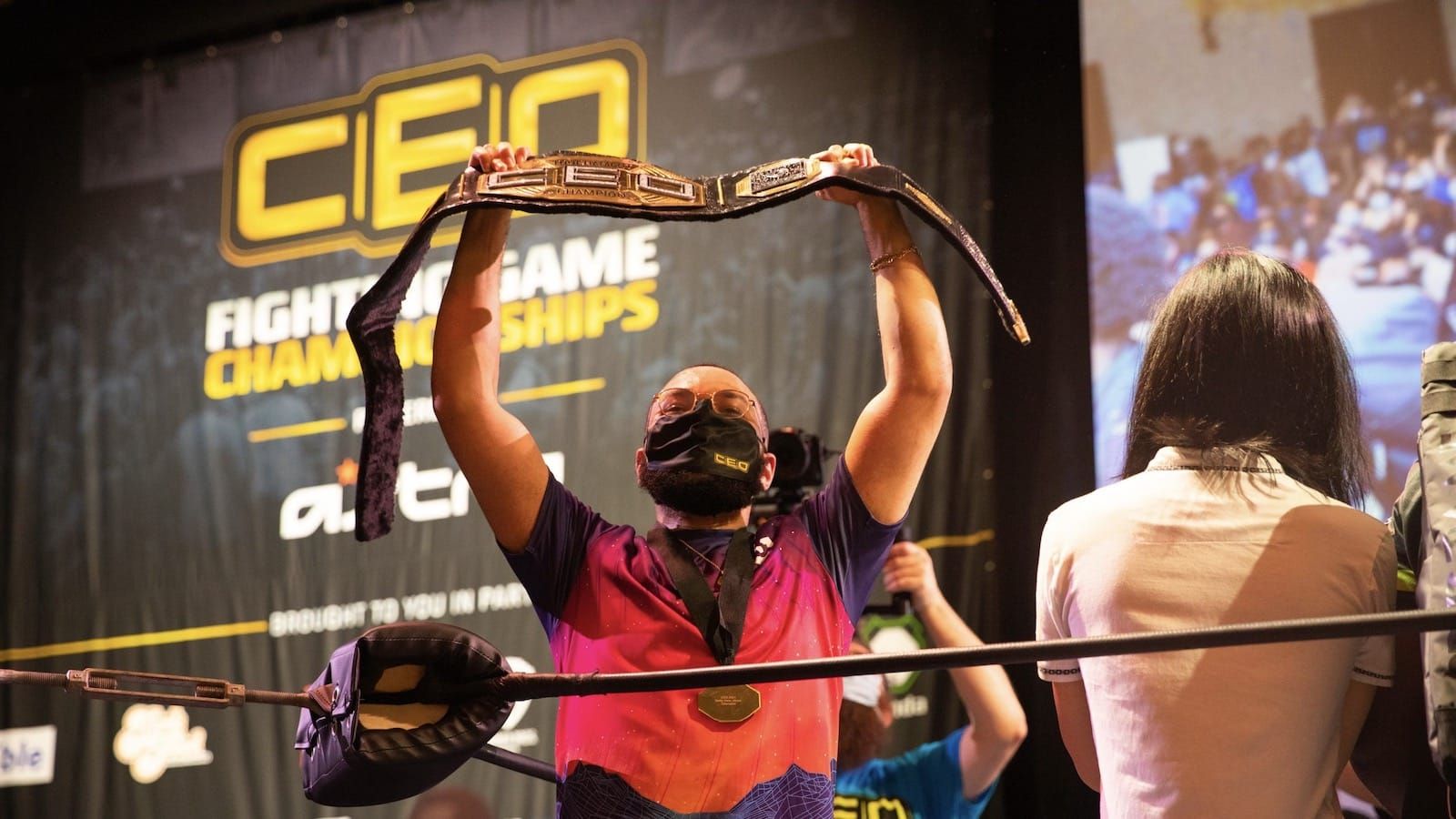 Fighting game player K7 lifts a championship belt above his head with name of tournament "CEO" in the background