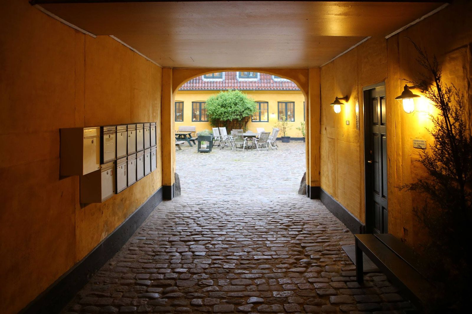 Arkitekturfoto af indgangsgang med gul stenvæg, koblingsledninger og udsyn til gård med møbler og træ