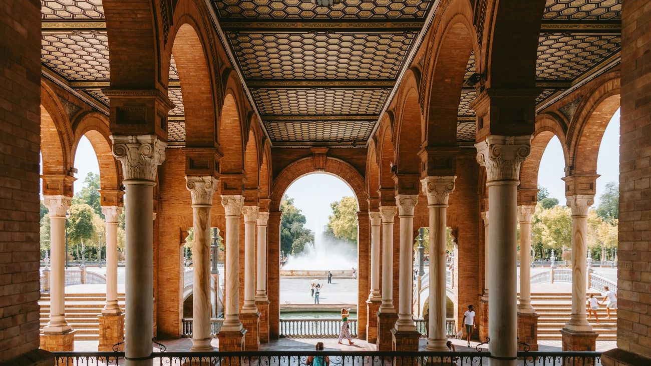 Brick arcade with marble columns framing a sunlit plaza and fountain
