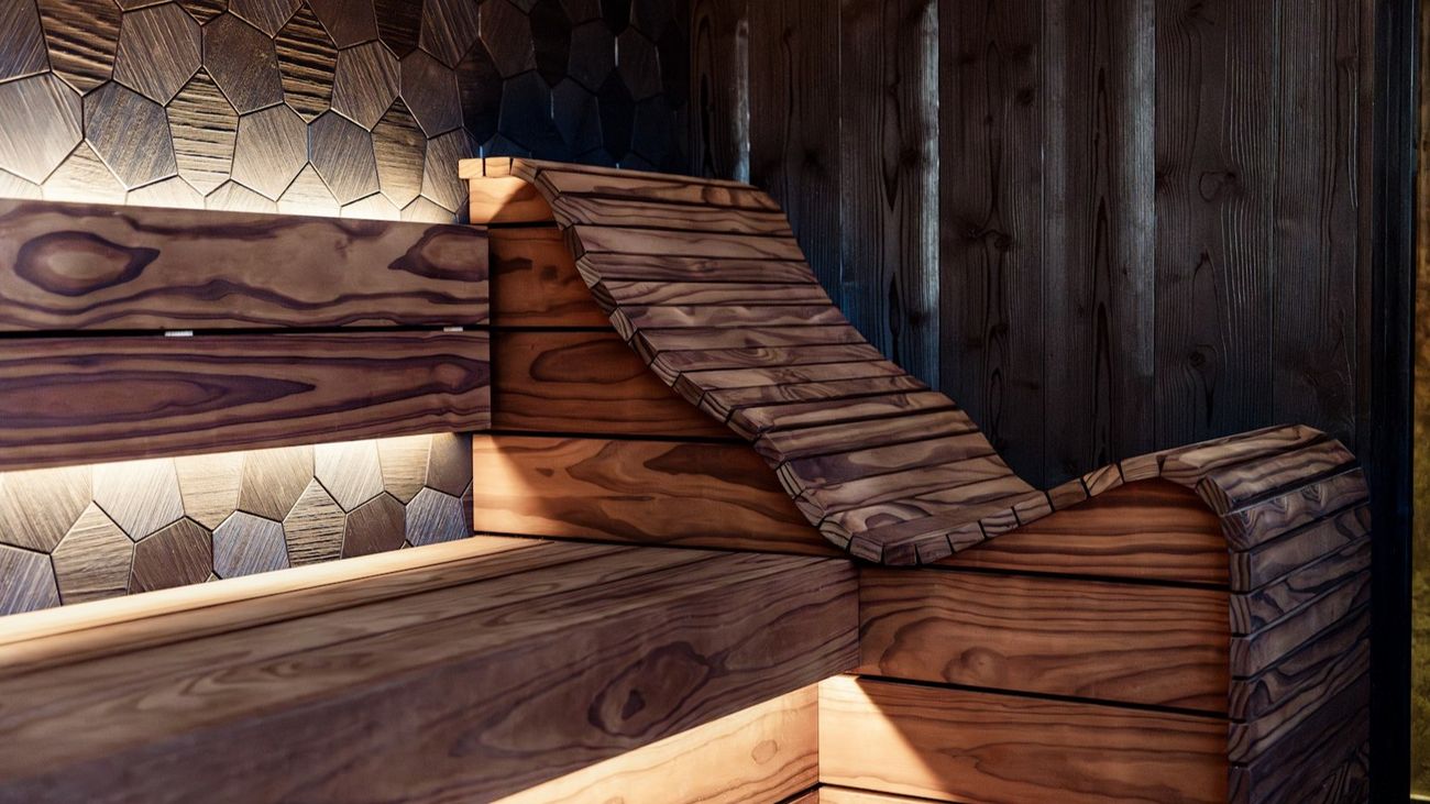 A curved wooden sauna lounger lit by warm amber light against a wall of hexagonal wood tiles.