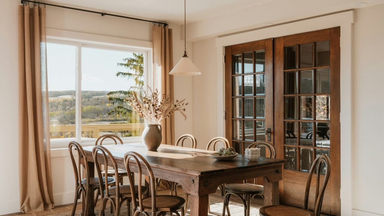 A sunlit dining room with a long wooden table, bentwood chairs, linen curtains and a view over hills.
