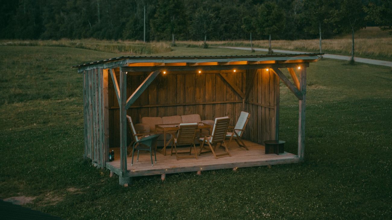 A wooden open-front shelter with string lights and outdoor chairs on a lawn at dusk